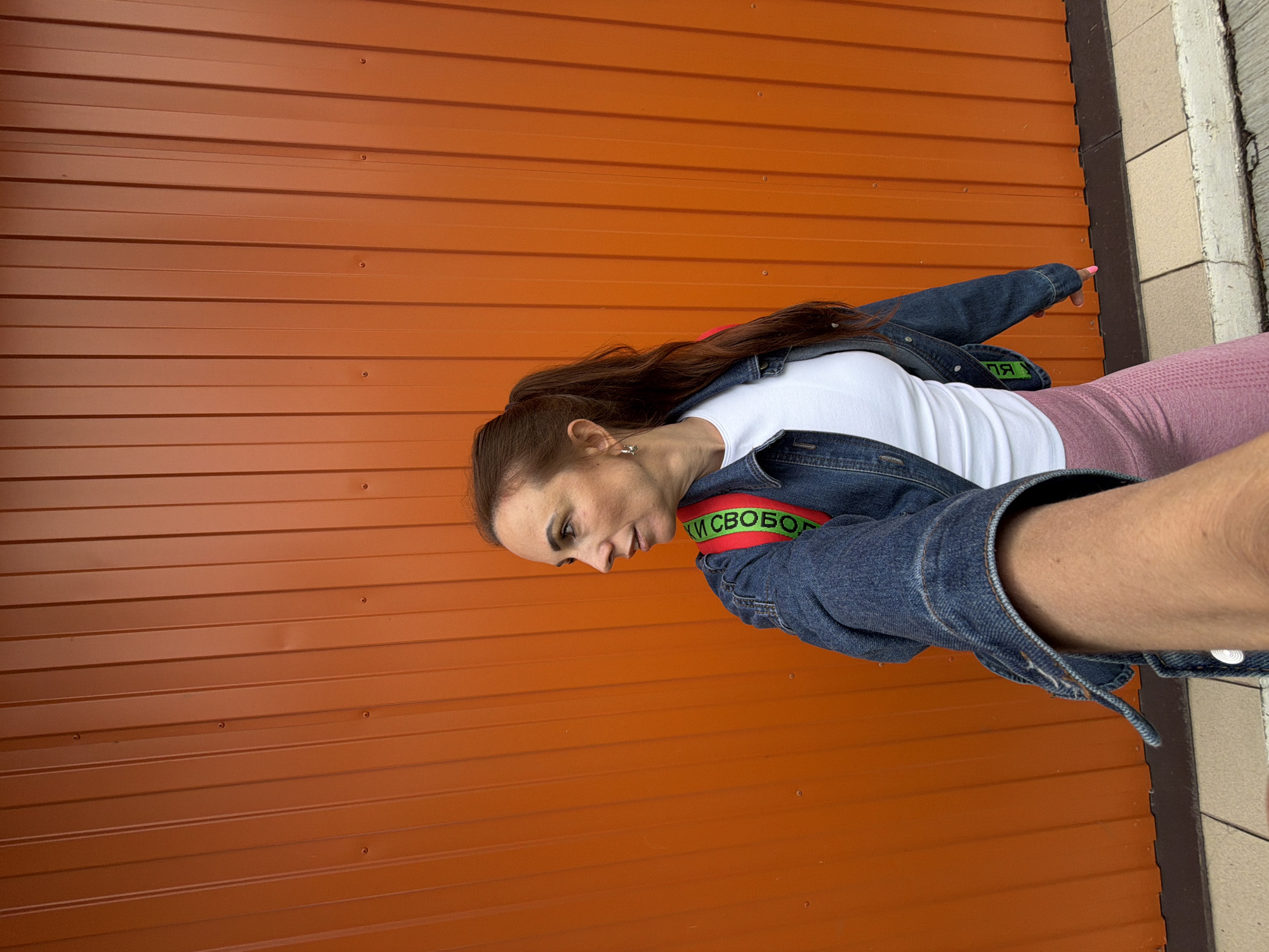 The image shows a woman with her hair in a ponytail, wearing a denim jacket over a white t-shirt and pink leggings. She is standing in front of an orange corrugated metal wall, likely outdoors in an urban or industrial area. A backpack strap with the word "СВОБОДА" (Freedom) in Russian is visible over her shoulder. The ground in front of the wall appears to be paved with tiles and concrete. The lighting suggests it is daytime. There is no specific indication of the city of Birobidzhan, Russia, within the image itself, other than what is stated in the prompt.