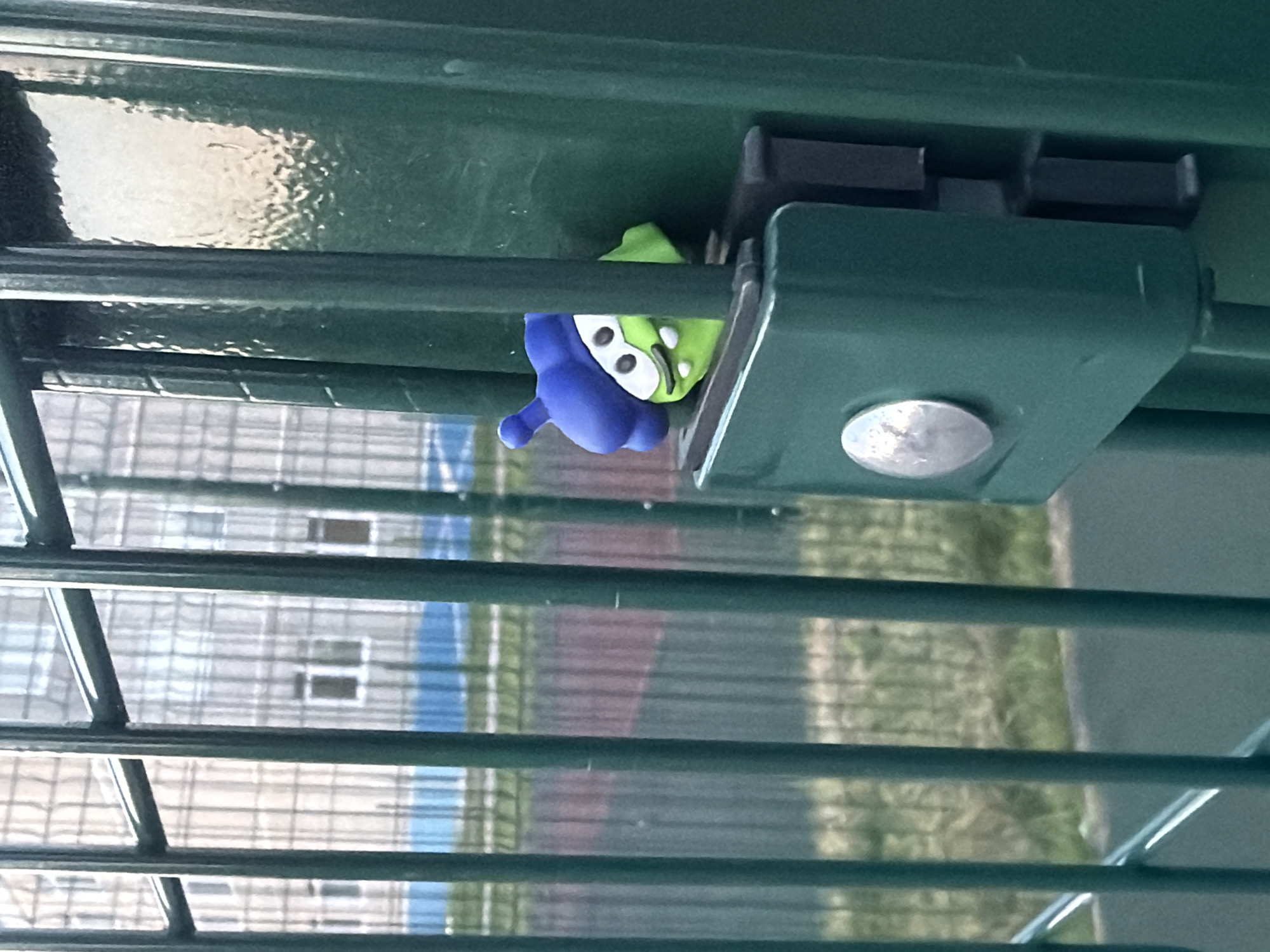 A close-up shot depicts a green metal structure, likely a fence, with parallel bars running horizontally across the frame. A small, stylized figurine resembling a green alien with a blue hood is clipped onto the fence's locking mechanism. The alien figurine is partially obscured by the locking mechanism. The background, visible through the fence's bars, shows a building or structure with multiple windows. The location of this image is Markovskii, Russia.