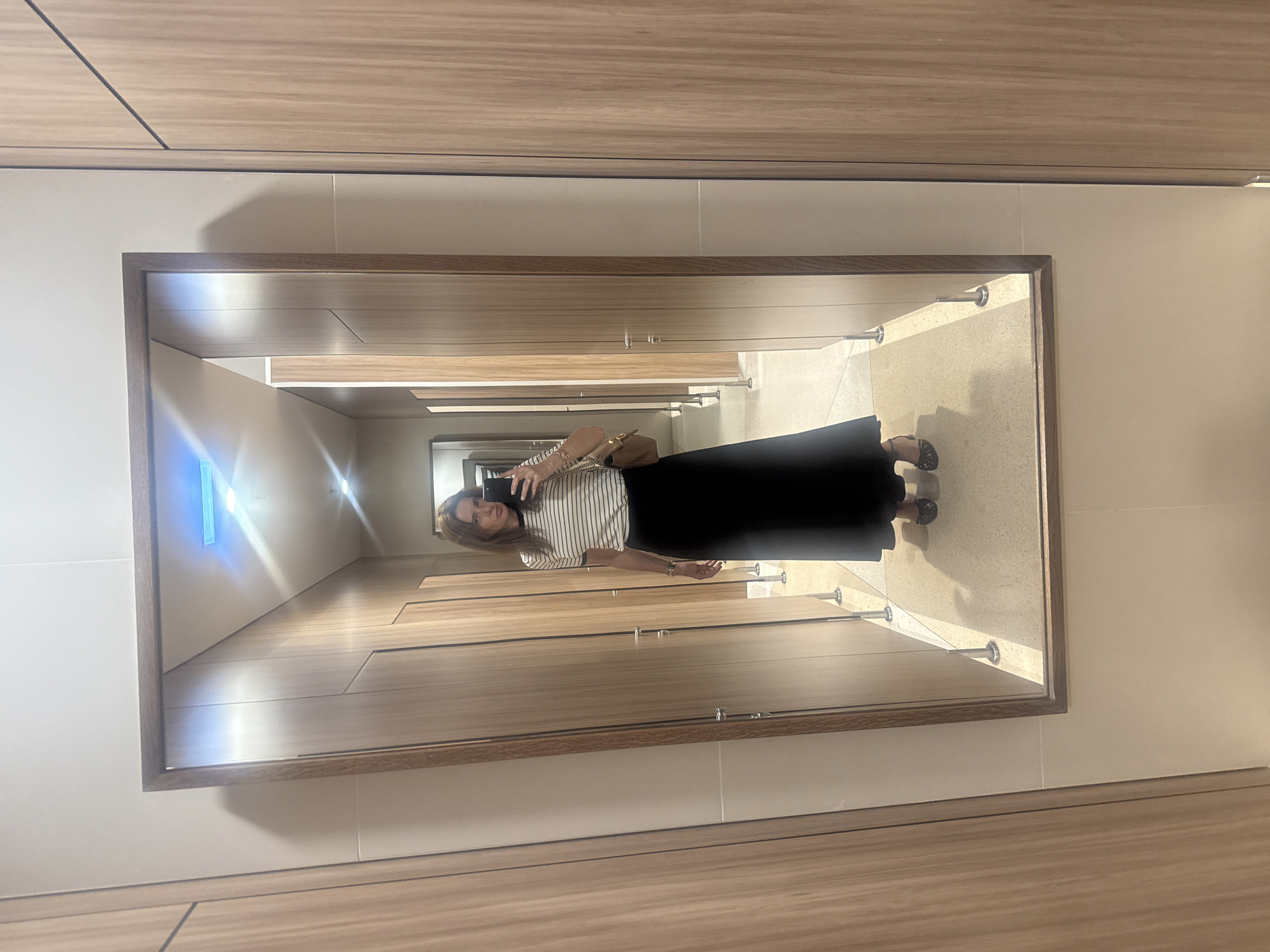The image depicts a woman reflected in a mirror in what appears to be a restroom. The woman is holding a phone and taking a picture of herself. She is wearing a striped shirt and a long black skirt. The restroom has multiple stalls, with wood-paneled walls and a light-colored tiled floor. There is no information in the image that indicates the location in the city of Torres Vedras, Portugal.