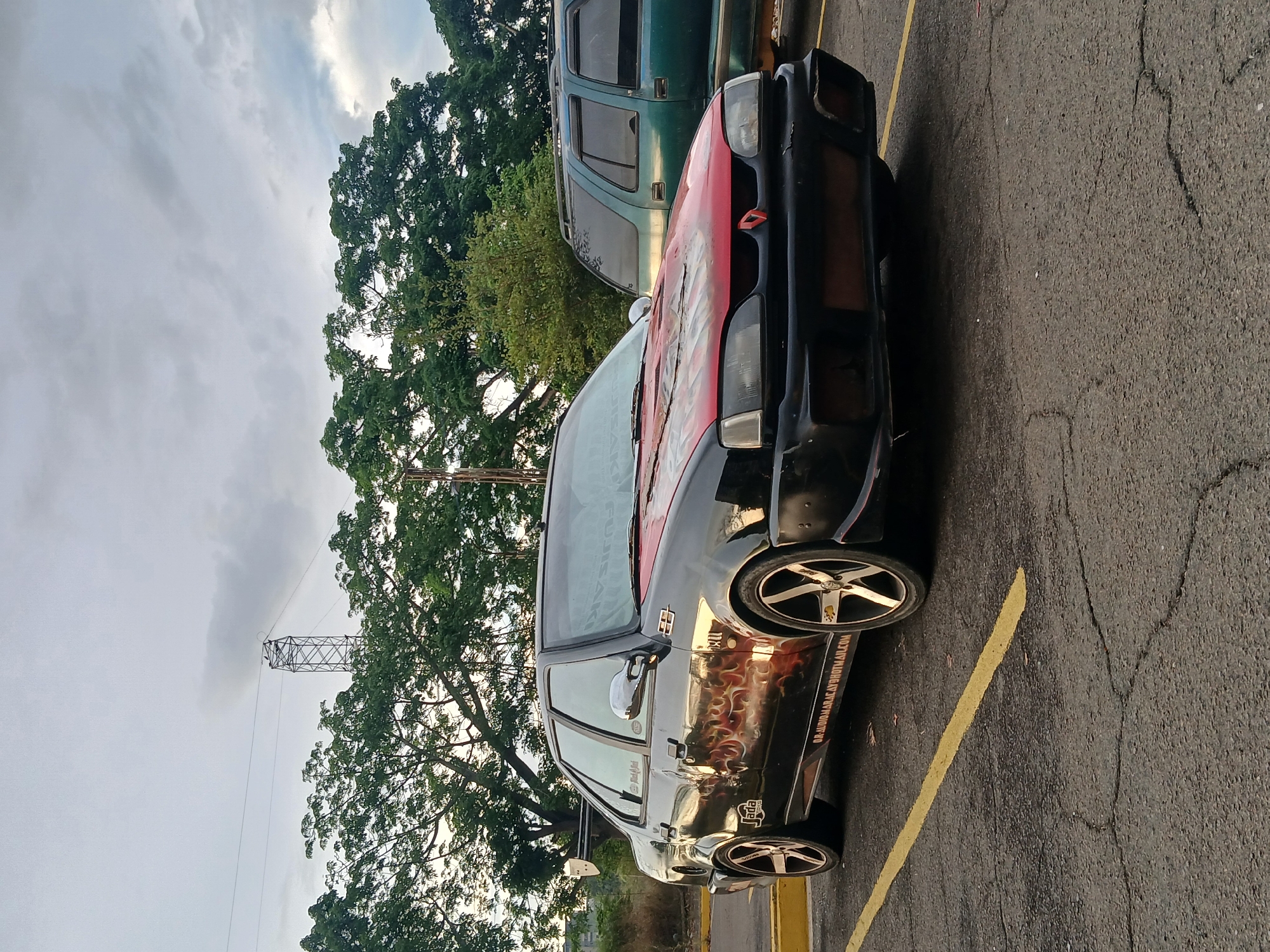 This media file presents an outdoor scene featuring two parked cars under an overcast sky. The primary subject is a black sedan, heavily customized, positioned at an angle on the right side of the frame. Its hood is red with some faded white or light-colored graphics, and its lower sides are adorned with an orange and red flame decal. Custom multi-spoke silver rims are fitted on the wheels. A prominent text "04-AUDIOMARACAY@HOTMAIL.COM" is visible on the car's side, suggesting contact information or an affiliation. A smaller sticker near the rear wheel reads "lada," indicating the car's make. The front bumper appears modified or has missing elements. Behind this car, a green SUV or truck is partially visible.
The vehicles are situated on a cracked asphalt surface with yellow painted parking lines. In the background, lush green trees with dense foliage dominate the left side of the frame, hinting at a natural or suburban environment. Further in the distance, a tall metal electricity pylon or tower stands against the sky. The sky itself is mostly cloudy, with some brighter patches suggesting a soft, diffused light, characteristic of an overcast day, possibly late afternoon or early morning. There is no explicit activity taking place, and no people are visible. The scene depicts a static moment within what appears to be a parking area in Ciudad Guayana, Venezuela.