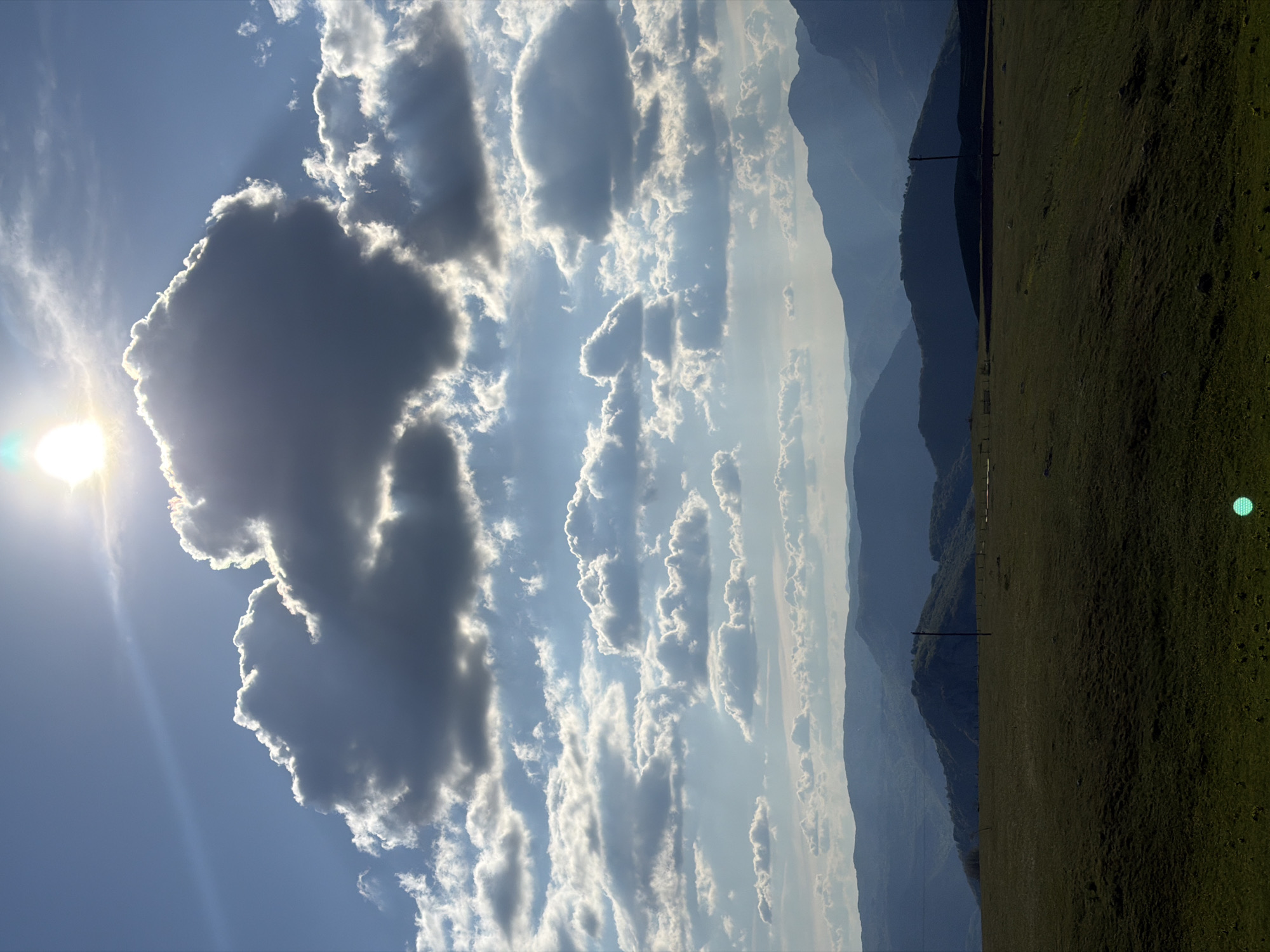 The image depicts a rural scene, presumably in or near Pišče, Montenegro, given the prompt's context. The sky dominates the view, featuring a mix of clear blue and large, dark cumulonimbus clouds illuminated by the sun. Sunlight streams through gaps in the clouds. A range of mountains is visible in the distance. The foreground is primarily a green field. Power lines run across the field, partially obscuring the mountains behind.