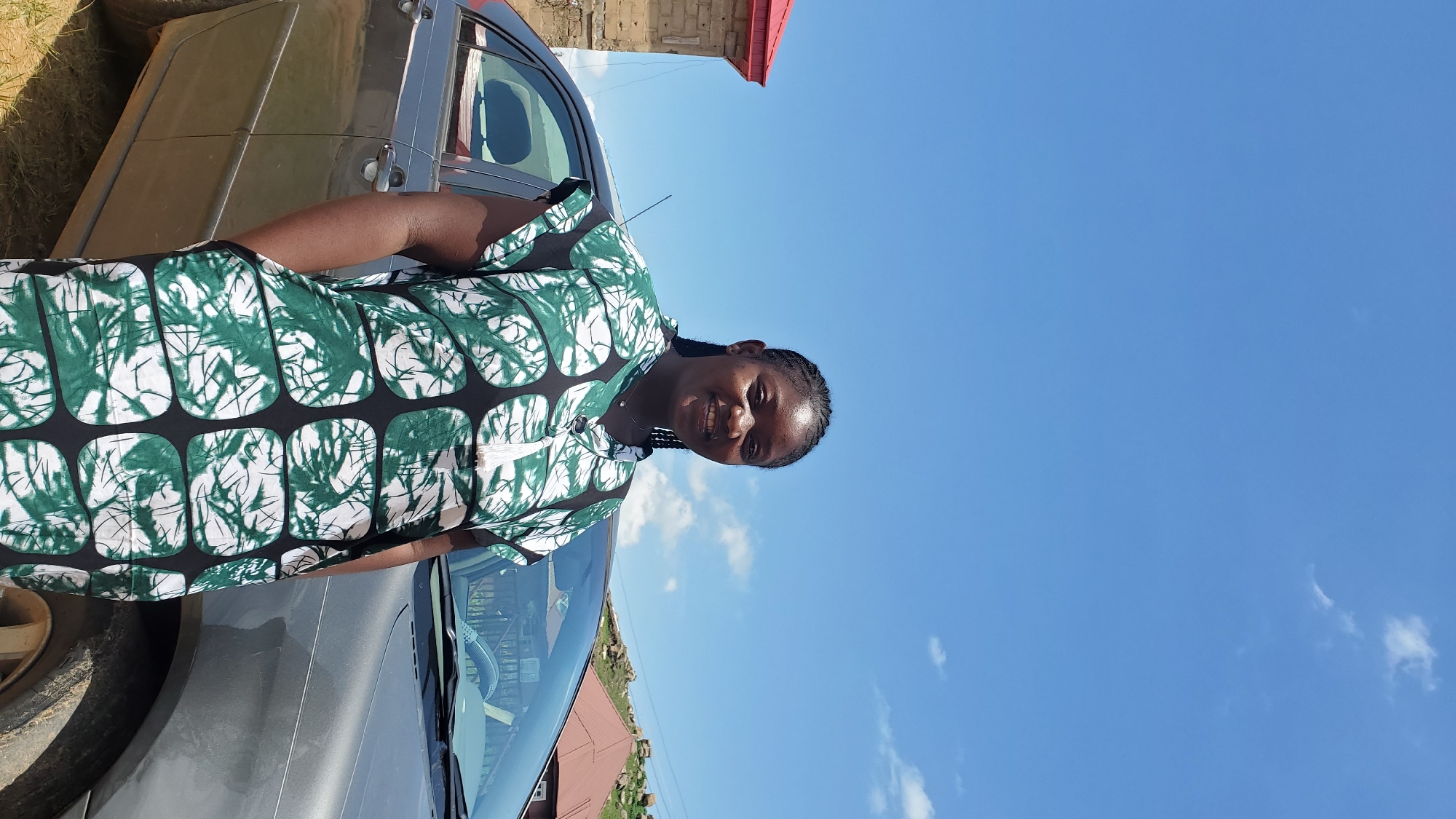 A full-body shot shows a young Black woman standing in front of a gray sedan with her left hand in her pocket. She has her right arm bent and resting on her hip. The woman has dark braided hair and is smiling at the camera. She is wearing a green and white patterned dress with a v-neck and a white tassel detail. The car is parked on a dirt surface. In the background, there is a brick building with a red roof on the left and a house with a red roof on the right. Hills with sparse vegetation are visible in the distance. The sky is bright blue with scattered white clouds. The scene appears to be in an outdoor, possibly rural, location in the city of Barakin Akawo, Nigeria.