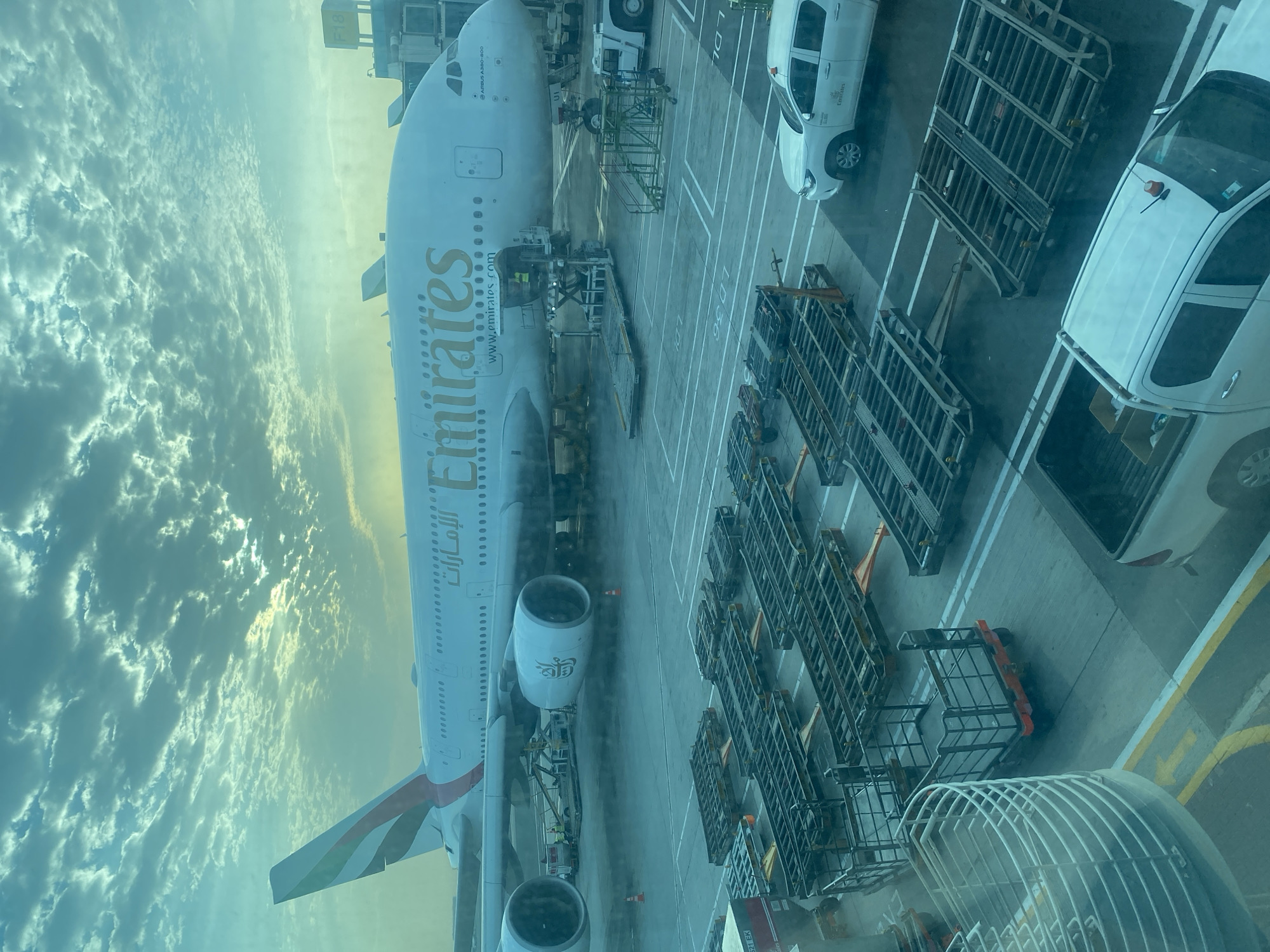 This image captures a large Emirates Airbus A380-800 aircraft parked at a gate on an airport tarmac in Dubai, United Arab Emirates, during either sunrise or sunset. The scene is viewed from an elevated perspective, likely from inside an airport terminal window.
The enormous aircraft is the central subject, featuring its distinct white livery with the gold "Emirates" logo in both English and Arabic script on its fuselage, along with "www.emirates.com". The designation "AIRBUS A380-800" is also visible near the cockpit. Its engines and a portion of its tail fin, adorned with the Emirates flag design, are also in view.
Around the aircraft, the tarmac is busy with ground support equipment and vehicles. Numerous metal baggage containers (ULDs) are arranged in rows, indicating baggage handling operations. Various airport service vehicles, including white vans (one with an Emirates logo) and utility trucks, are parked on the tarmac. Faint ground markings, including "LD 36," are visible. A jet bridge, identified by the text "F18," connects to the aircraft's upper deck, suggesting passenger boarding or disembarking.
The sky on the left side of the frame is filled with dynamic, scattered clouds, illuminated by warm, soft light, characteristic of the golden hour. This lighting casts a warm glow over the entire scene, creating a vibrant backdrop to the airport activity. The image conveys the organized operations surrounding a large commercial airliner at a major international hub.