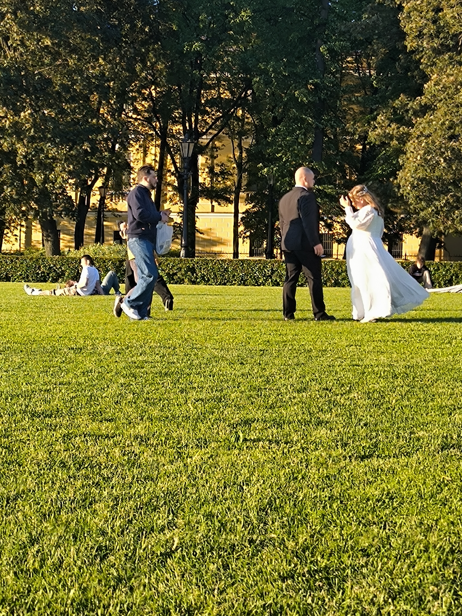 In a sunlit grassy park, a wedding party is captured mid-event. A bride in a flowing white gown and a groom in a dark suit stand facing each other, the bride adjusting her hair. To their left, a man in a dark jacket and jeans walks away, carrying a white bag. In the background, two people recline on the grass, appearing relaxed. The scene is set against a backdrop of lush green trees and a yellow building.