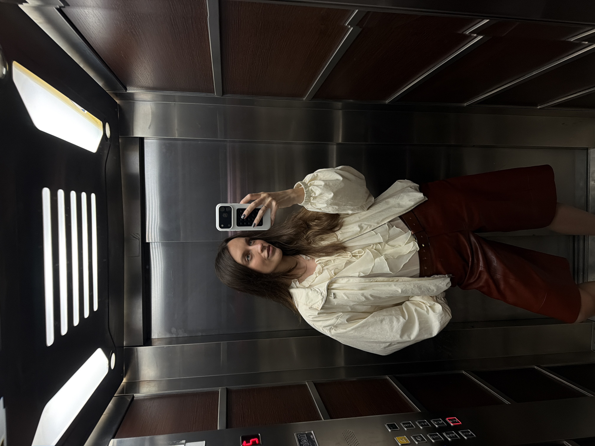 A young woman is captured in a mirror selfie taken inside an elevator, looking directly at the camera with a slight smile. She has long, dark brown hair and is wearing an off-white, high-necked top or jacket with ruffled details and puffed sleeves, paired with dark reddish-brown shorts or a skirt, possibly made of leather. Her left hand, adorned with a ring on the ring finger and dark nail polish, holds a smartphone with a white case, reflecting her image.
The elevator interior features a dark brown wood-grain ceiling with silver trim. The walls are a mix of reflective stainless steel panels and dark, possibly black, textured panels with vertical white strip lights on the left. A control panel is visible at the bottom of the reflected image, displaying a red "5" on a digital screen, indicating the current floor. Several silver-colored square buttons with numbers (1-9, 0) and symbols are also present on the panel, alongside some unreadable text. The overall lighting is artificial and bright inside the confined space. The setting is likely a modern building, situated in Budva, Montenegro, as per the context.