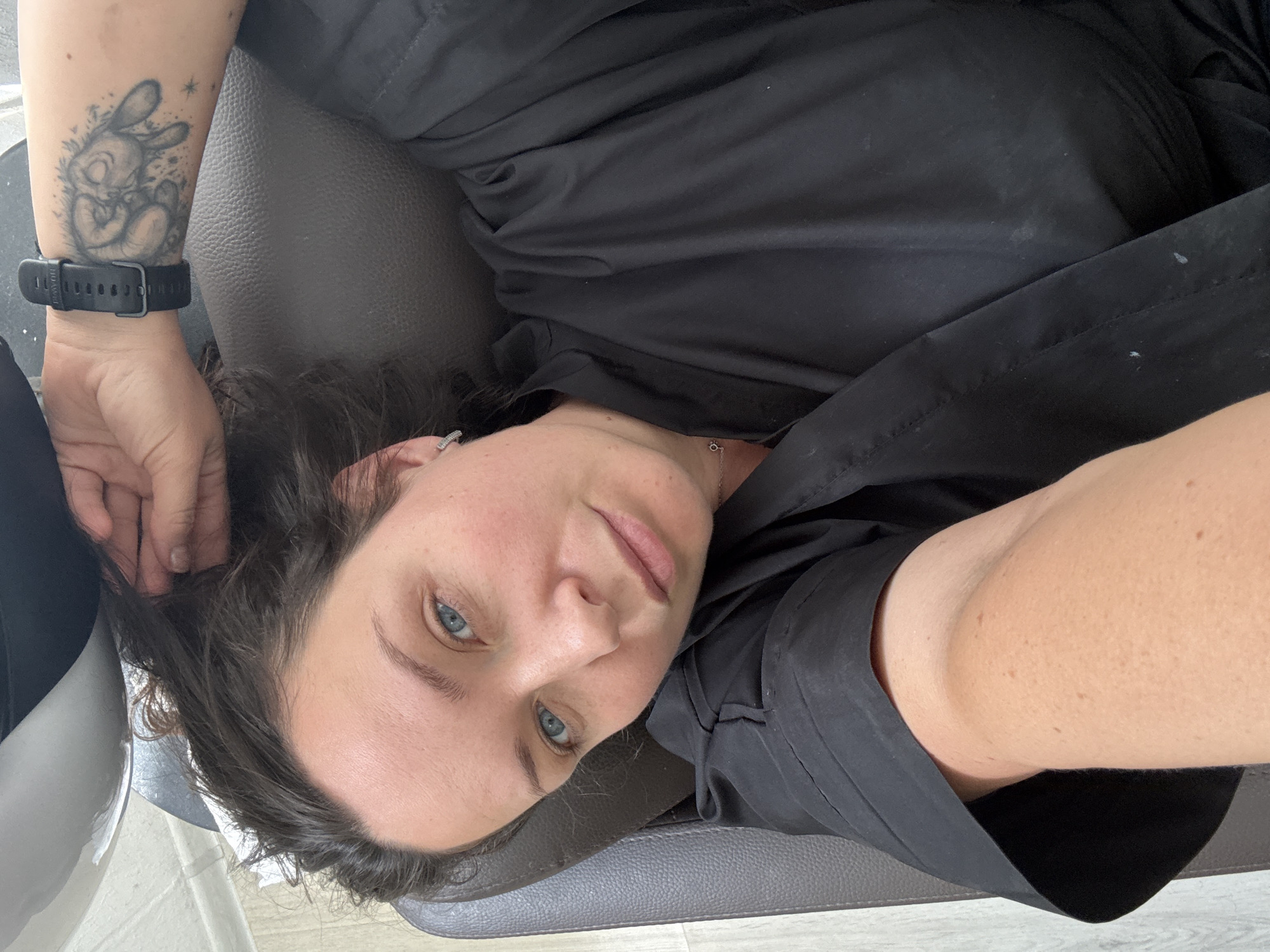 A woman with blue eyes and dark hair lies on a brown leather chair, looking at the camera. She is wearing a black robe and a black watch on her left wrist. Her left arm is extended, holding her hair back. A tattoo of a hand giving the peace sign is visible on her left forearm. The scene appears to be indoors, possibly in a salon or spa, with a light-colored wall in the background. There are no specific landmarks or indications of the city of Chaikovsky, Russia.