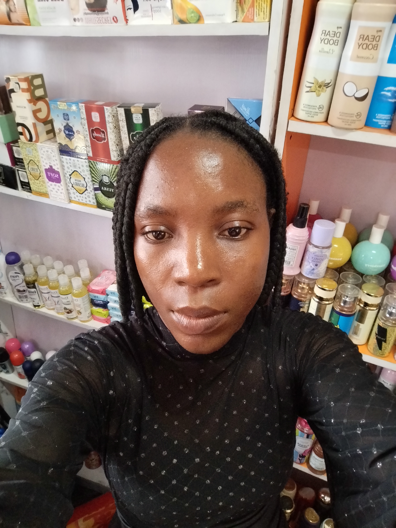 A medium shot from chest-up shows a young Black woman with dark skin and braided hair styled into two pigtails that fall over her shoulders. She is wearing a sheer, black, long-sleeved top with a subtle sparkly diamond pattern. Her face is angled to the left, with her eyes looking directly at the camera. Her lips are slightly parted, and her skin appears dewy. She is positioned in front of shelves stocked with various cosmetic and personal care products. On the shelves behind her are rows of bottles and boxes of perfumes, lotions, and soaps. The shelves appear to be in a retail setting. The lighting is bright, suggesting an indoor environment, possibly a shop in Ugbowo, Nigeria.