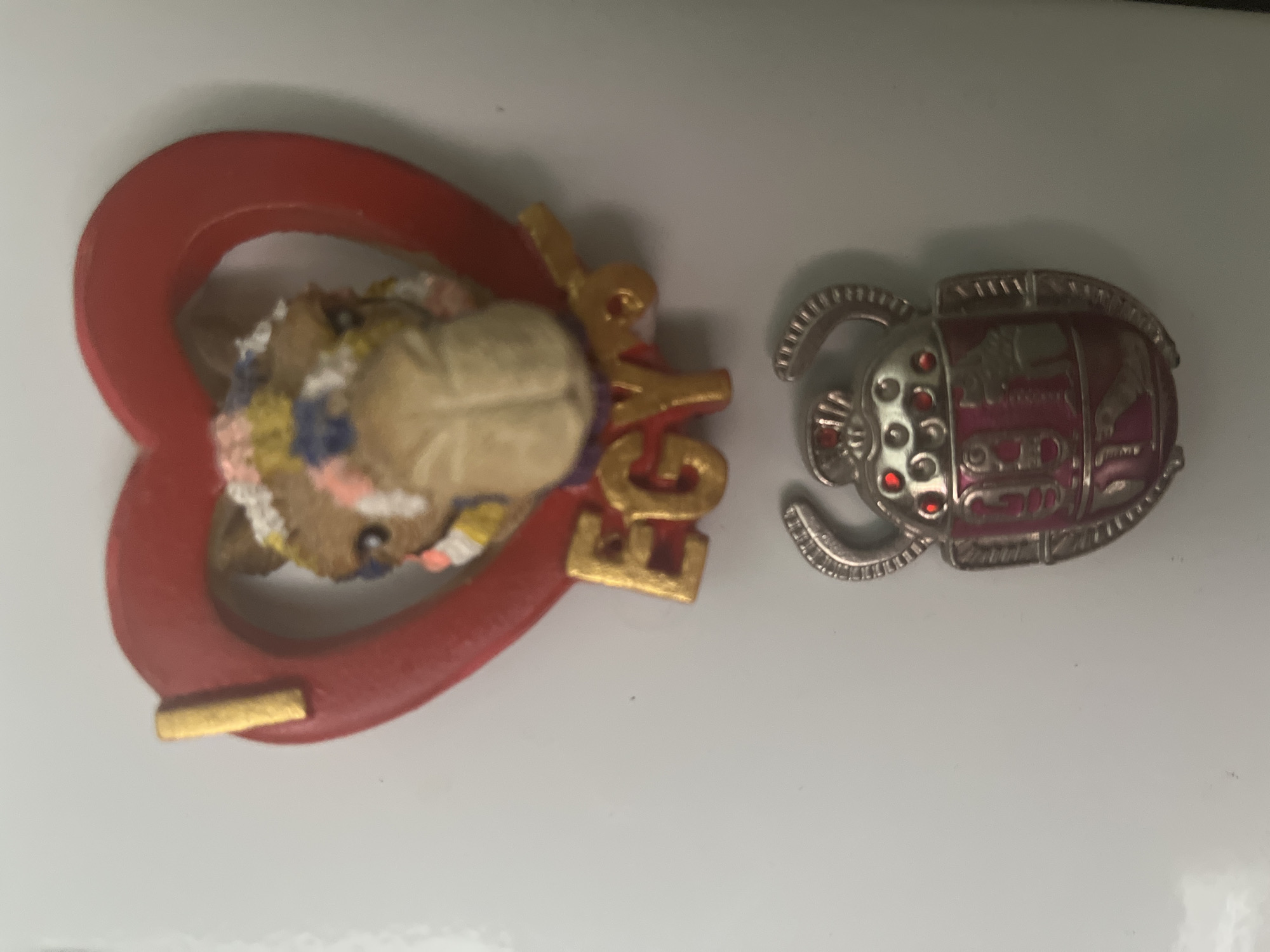 The image shows two Egyptian-themed magnets on a white surface. The top magnet is a red heart with a camel's head in the center, adorned with colorful beads. The word "EGYPT" is written in gold lettering below the heart. The bottom magnet is a silver scarab beetle with red gem accents and intricate details. The overall scene is well-lit and the objects are in focus. The location is not specified in the image and therefore cannot be determined.