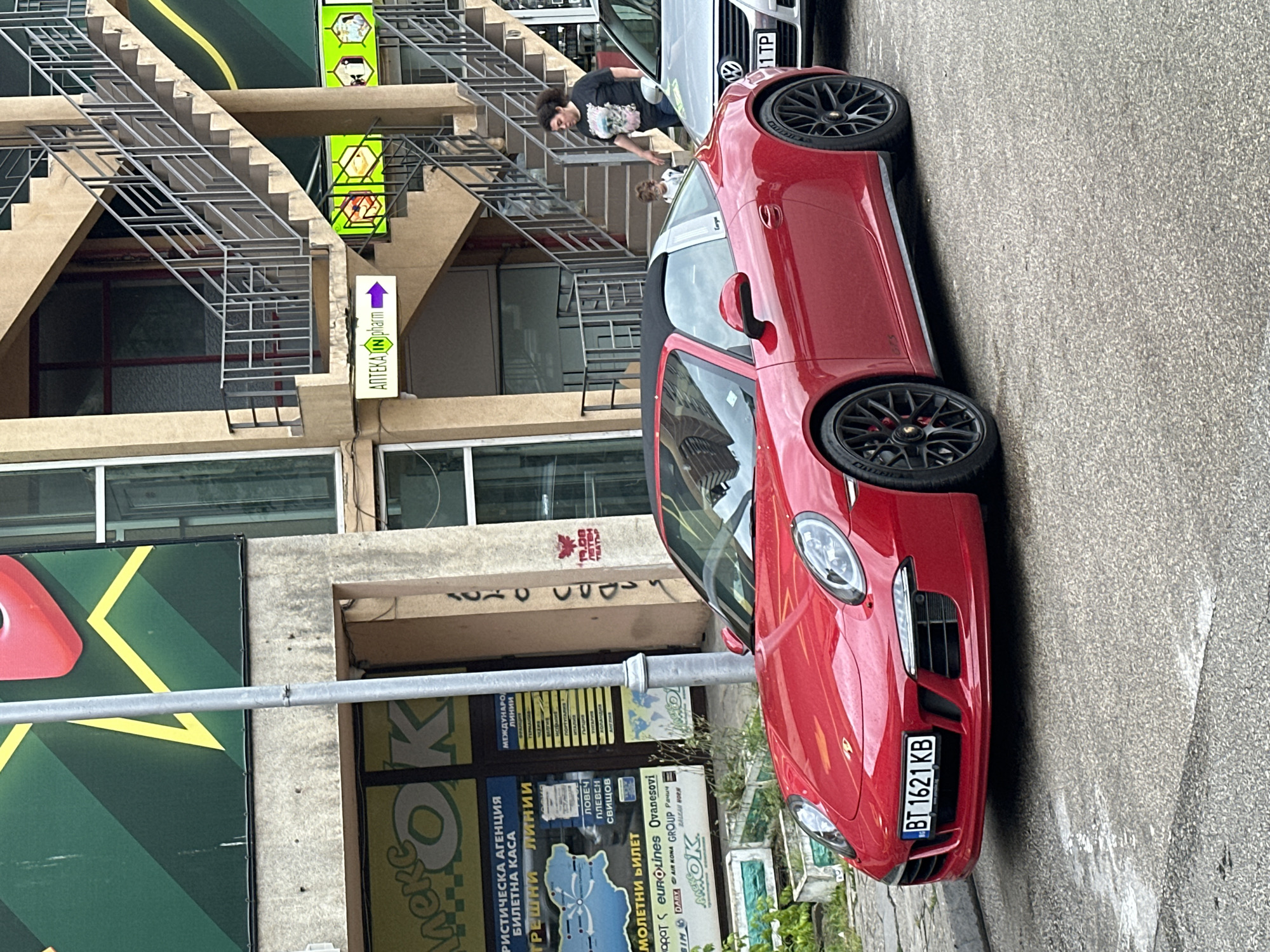 A red Porsche 911 Targa 4 GTS is parked on a street in Veliko Tarnovo, Bulgaria. The car is facing slightly to the left, with its front bumper visible. The license plate reads "BT 1621 KB". The car is parked in front of a building with an outdoor staircase. A woman and a small child are standing on the staircase, and a silver Volkswagen car is parked next to the Porsche. The building has several shops on the ground floor, including a pharmacy with a sign that says "ΑΠТЕКА IN pharm" and a directional arrow. There are also signs for a travel agency with routes plotted on a map of Bulgaria and international bus lines. The street is paved with asphalt and appears to be wet. The overall environment suggests a typical urban street scene in Bulgaria.