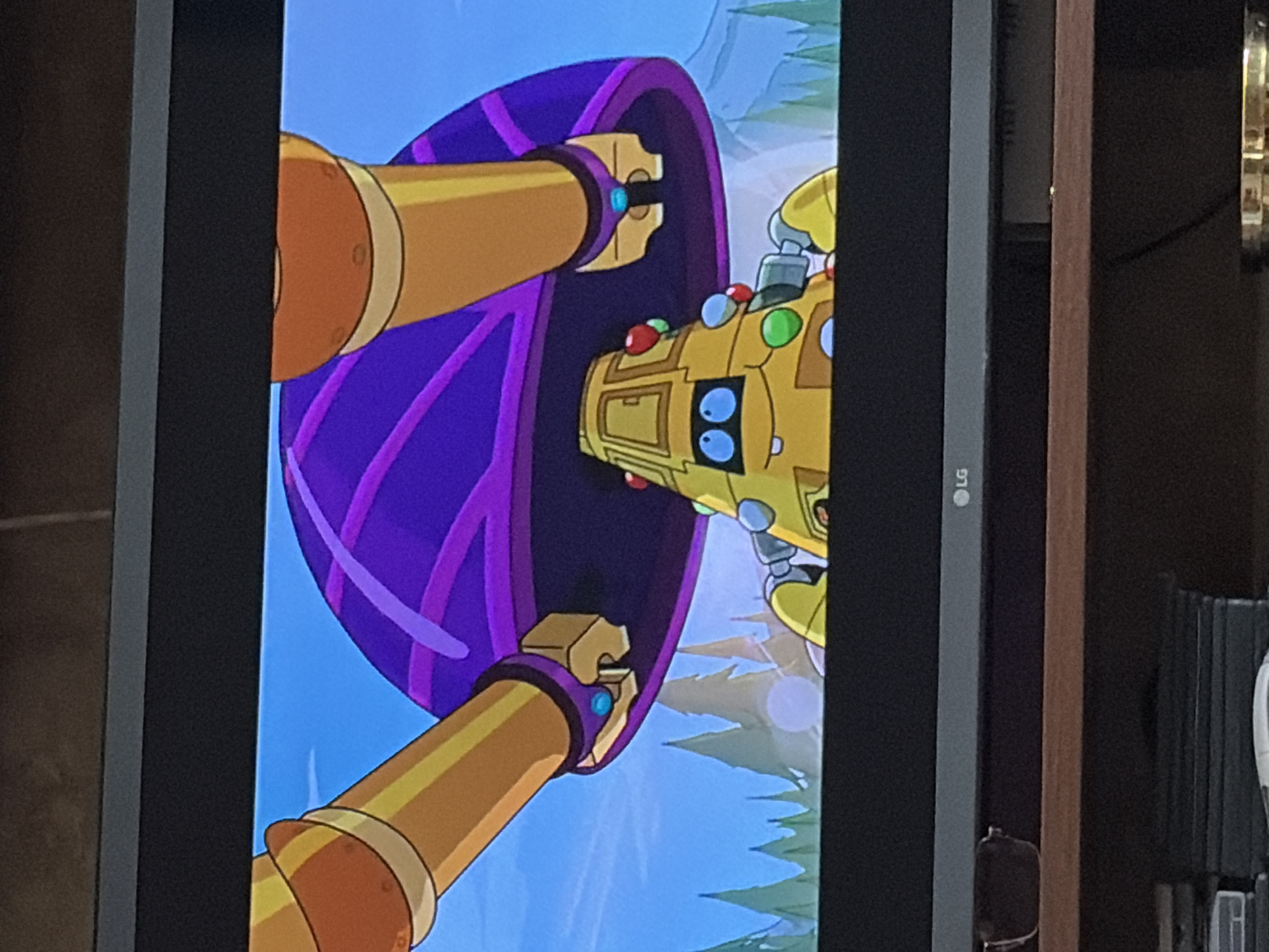 The image shows a close-up shot of a cartoon robot with a yellow, conical body adorned with colorful circular embellishments and a simple face with blue eyes and a small smile. The robot appears to be situated outdoors, with a background of stylized green trees and a light blue sky with faint clouds. Above the robot, a large, purple, sombrero-like object is visible, supported by two thick, golden, cylindrical arms that extend from the top of the frame. The scene is captured on a television screen, with the bottom portion of the TV frame and a shelf beneath it visible in the foreground. The overall impression is a still frame from an animated program.