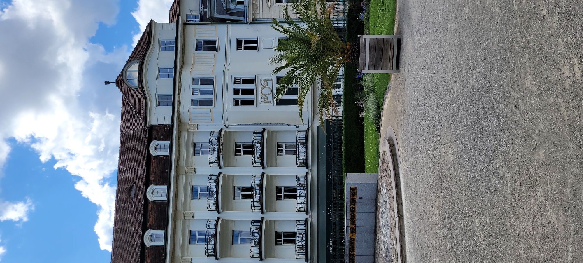 The image captures a scene in Baden, Austria, featuring a multi-story building with a light-colored facade and a dark tiled roof. The building exhibits architectural elements such as dormer windows on the roof and multiple balconies with wrought-iron railings. The balconies are furnished with small tables and chairs. Below the balconies, a green awning with repeating text that reads "HOTEL HERZOGHOF" is visible. In the foreground, there is a paved area leading to a circular stone structure with a curved stone border. Beyond this, a manicured green lawn is present, with a palm tree in a wooden planter box on the right side of the frame. The sky is blue with scattered white clouds. The scene is static, with no people or animals visible.