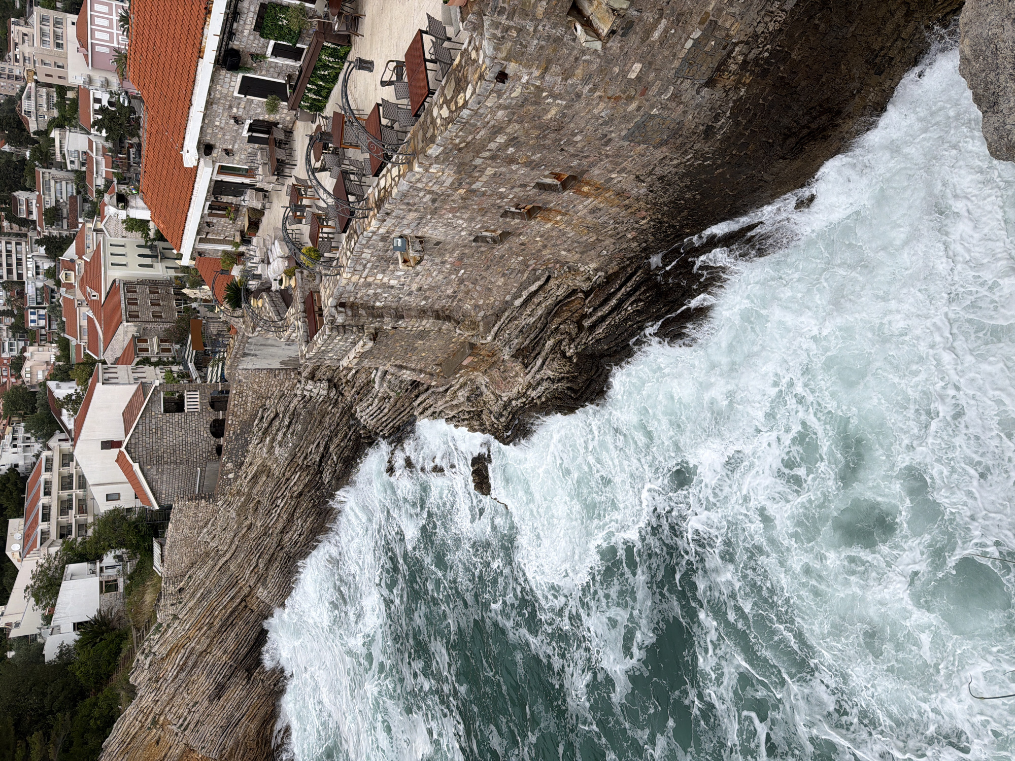 An aerial view of a coastal area in Petrovac centar, Montenegro. On the left side of the image are buildings with red tiled roofs nestled closely together, and further to the right is a restaurant or terrace area with tables and chairs. The rocky coastline, made of layered stone, borders the turbulent sea where waves crash against the rocks. The ocean water is a mix of white seafoam and a dark teal hue.