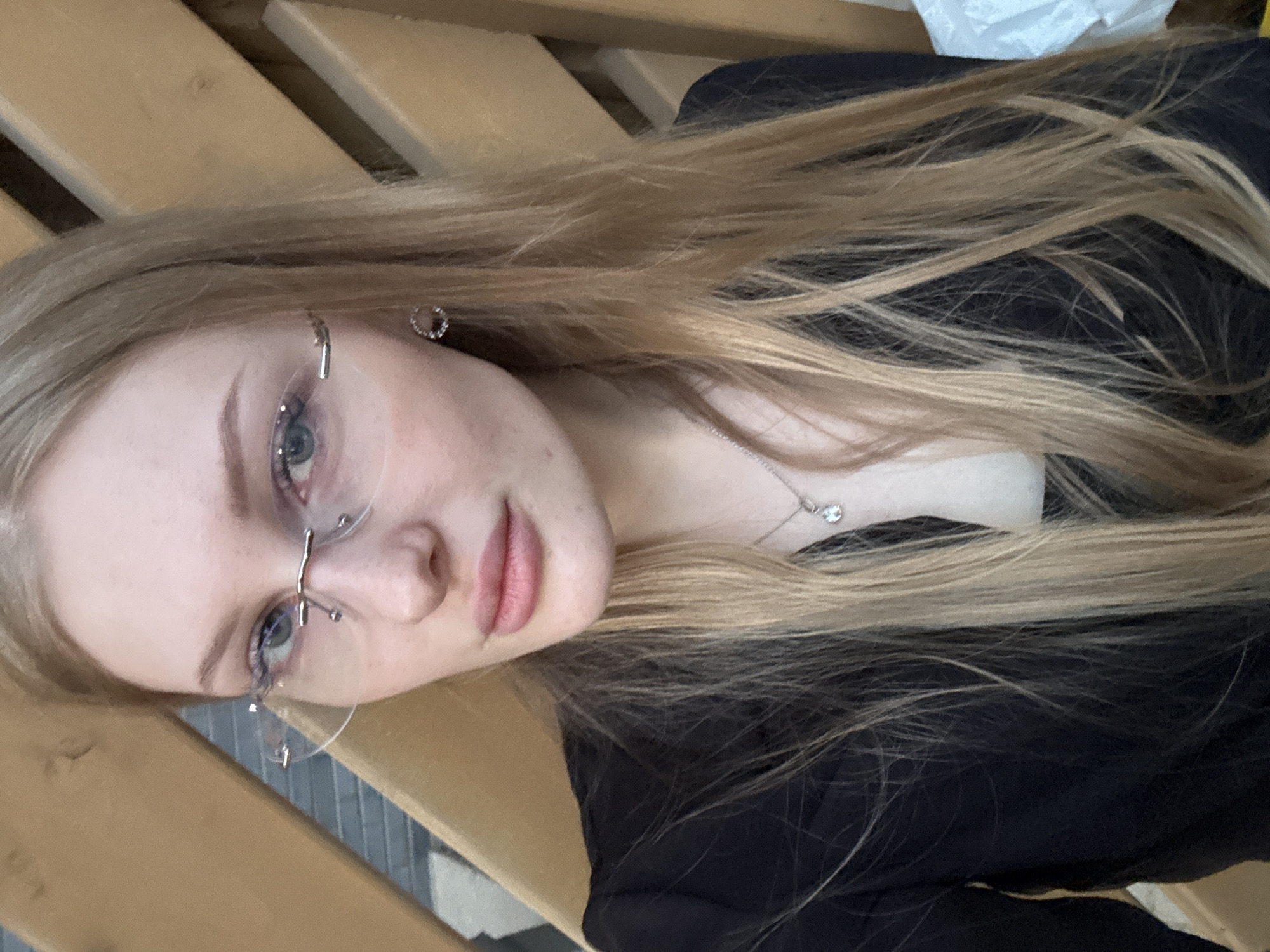 A blonde woman with glasses is photographed indoors. She is wearing a black top and a silver necklace. She has light skin and light-colored eyes. She is likely in Perm, Russia. The background includes wooden surfaces.