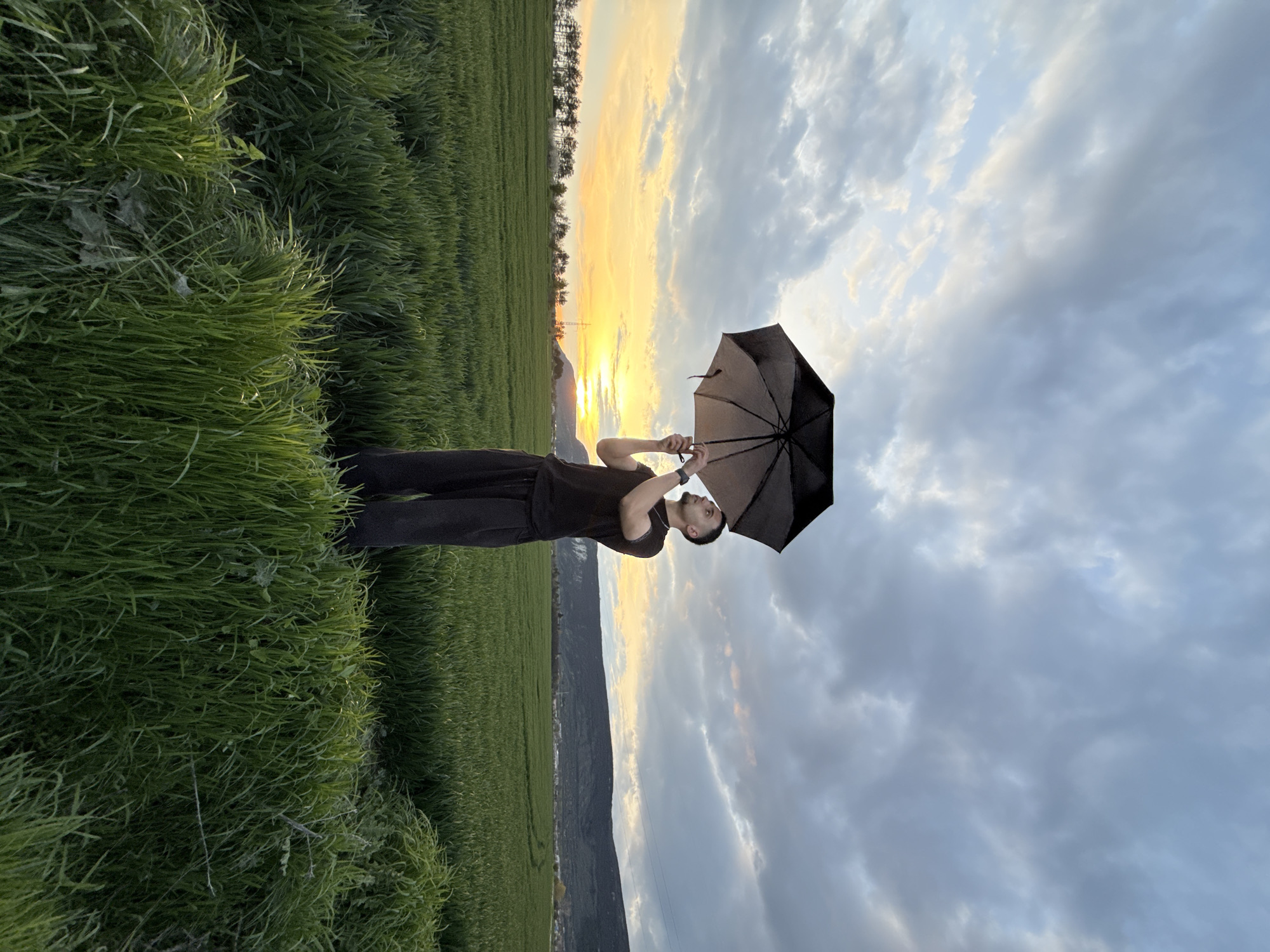 The image shows a man standing in a field with an open umbrella, against a backdrop of a sky with a sunset glow. The field is filled with green grass. In the distance, there are hints of a mountain range and possibly a city skyline. The man is wearing dark clothing. He is holding the umbrella above his head. The sky has a mix of clouds and clear areas. Given the prompt's request for location information in Shumen, Bulgaria, and using general knowledge, the environment suggests a location outside of the city center, potentially in the surrounding rural areas. It is impossible to give more details without exact geographical location information.