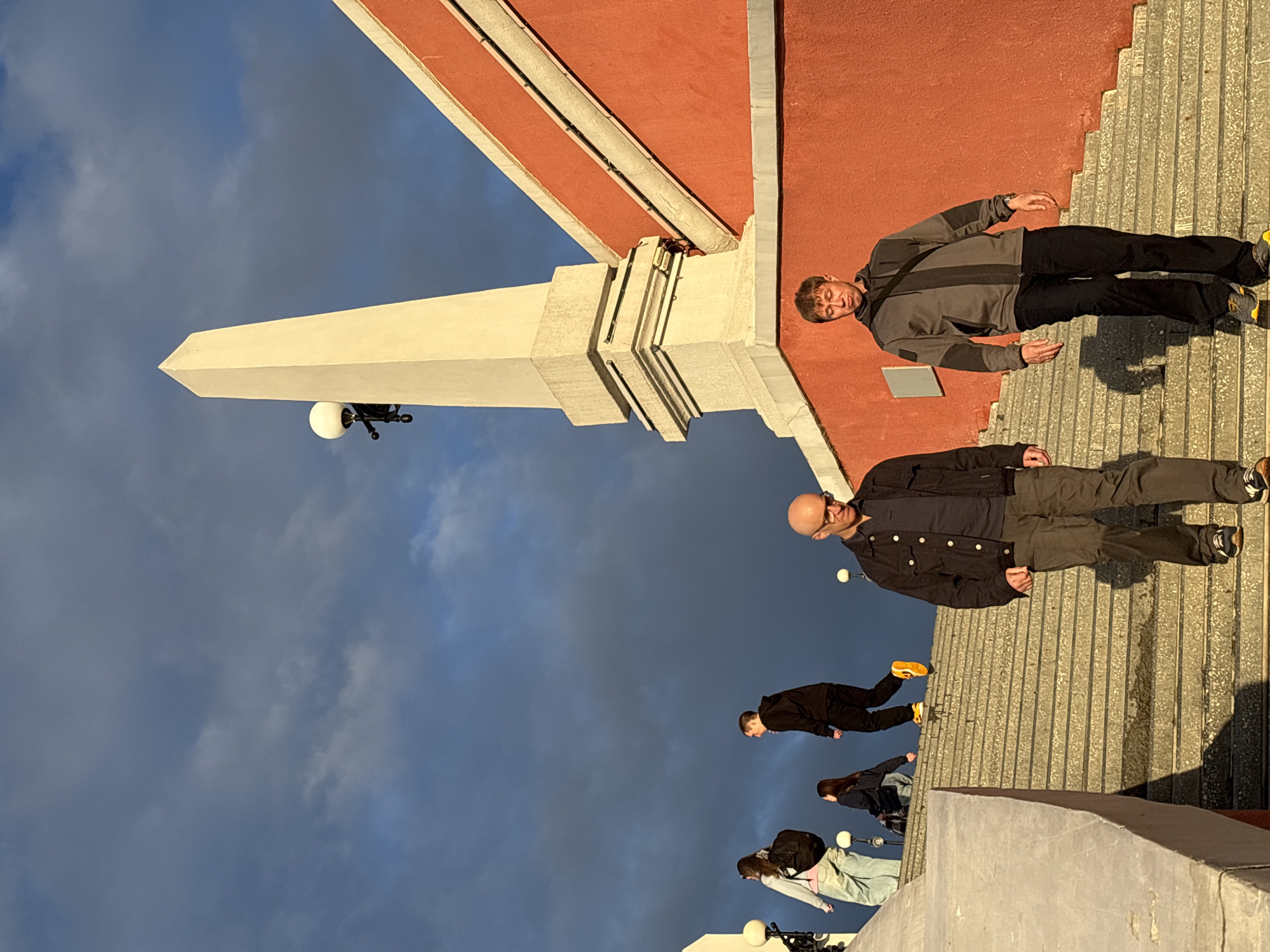 The image captures a scene on a wide, stone staircase in Nizhny Novgorod, Russia, with a striking red building and a white obelisk against a cloudy sky.
In the foreground, two men are walking down the stairs. The man on the right, wearing a grey hooded jacket and black pants, has his eyes closed and his head down. A black strap is slung across his chest. The man to his left, wearing a dark jacket and grey pants, is looking forward, with his hands at his sides.
Further up the stairs, several other individuals are visible. A person in a black hoodie and dark pants is walking up, with bright yellow shoes. To their left, a woman in a light-colored hoodie and light blue jeans, carrying a black backpack, is also ascending. Another woman, in a dark jacket, is walking up between the two.
The staircase is made of concrete or stone and is well-lit by the sun, casting shadows. The red building forms a vibrant backdrop to the scene, with its architectural details visible, including a light fixture on the obelisk. The sky is dramatic, with dark, heavy clouds suggesting impending weather. The overall impression is a moment captured in a public space in Nizhny Novgorod.