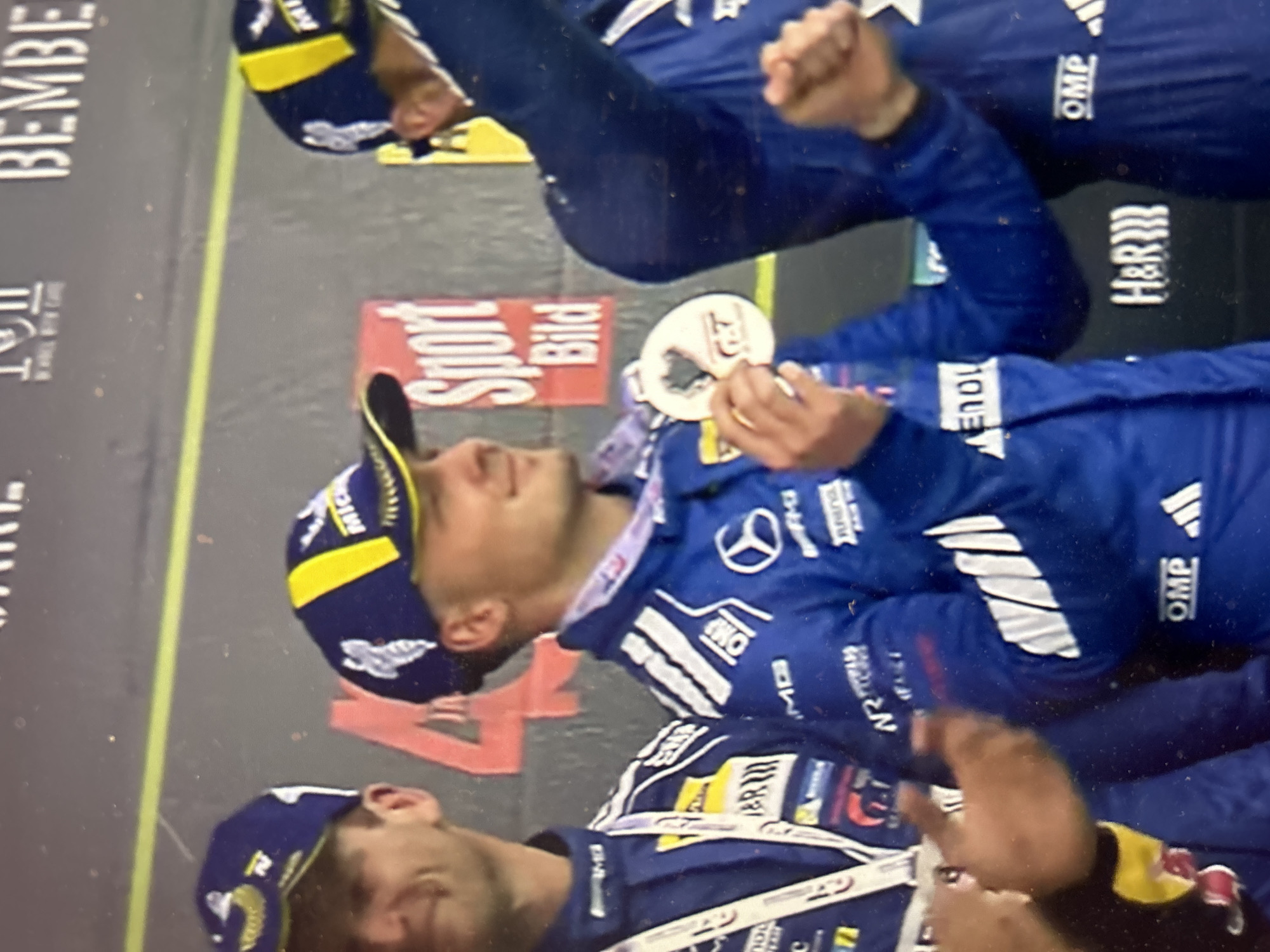 The image depicts three men wearing blue racing suits and caps with yellow accents. They appear to be on a stage or platform, possibly celebrating a victory or achievement. Each man wears a medal around their neck. Logos of various sponsors are visible on their clothing, including "AMG," "Mercedes-Benz," "H&R," "OMP," and "Michelin." One man is holding a white circular object with the number "24." A sign with "Sport Bild" is visible in the background. Other signs display the word "BEMBE" and "Arena." There is no clear indication that the location is in Sofia, Bulgaria.