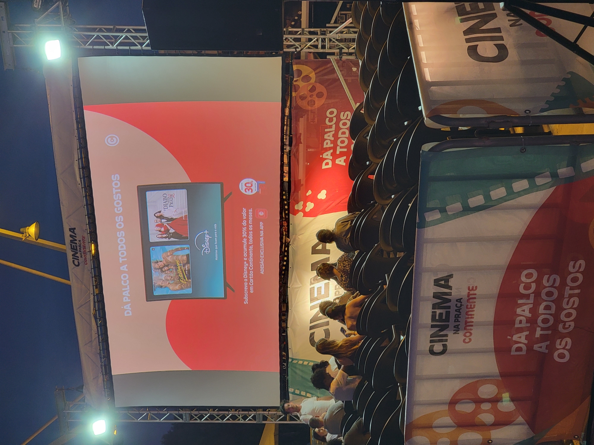 The image depicts an outdoor cinema setup at night in Quarteira, Portugal, specifically at the Praça do Continenete. A large projection screen displays a promotional advertisement for Disney+, featuring movie posters and text in Portuguese. The screen is illuminated by stage lights, casting a bright glow on the surrounding area. In front of the screen, rows of empty dark seats are arranged, suggesting an outdoor cinema or event space. Banners with "CINEMA NA PRAÇA CONTINENTE" and "DÁ PALCO A TODOS OS GOSTOS" (Cinema in the Square Continent, From Stage to All Tastes) are visible, further identifying the location and event. Several people are visible in the mid-ground, some standing and some seated, facing away from the camera and towards the screen. The overall atmosphere is that of a community event held in an open public space.