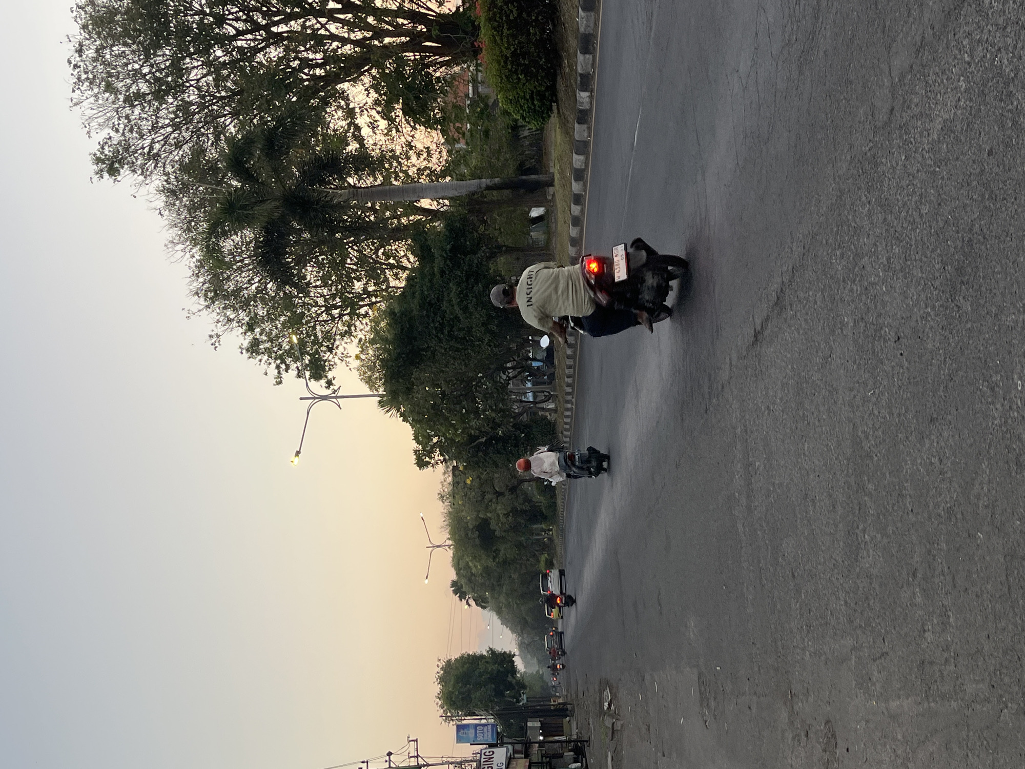 The image depicts a street scene in Ngaban, Indonesia, at either sunrise or sunset, as indicated by the soft, warm lighting and clear sky. The foreground is dominated by an asphalt road with visible cracks and some debris. Two motorcycles are the primary subjects.
On the right side of the road, a person wearing a light-colored t-shirt with the word "INSIGHT" printed on the back is riding a dark-colored motorcycle. The rider is wearing a baseball cap and dark pants. The motorcycle's license plate is partially visible and appears to read "W 4188 NGO". The brake light of this motorcycle is illuminated.
Further down the road, on the left side of the lane, another person is riding a dark-colored motorcycle. This rider is wearing a bright orange helmet and a light-colored jacket or shirt.
Several other vehicles, including cars and motorcycles, are visible in the distance, suggesting moderate traffic. The left side of the road features a sidewalk with some shops or buildings, including one with signage that includes the word "GING" and another with "SOTO". Power lines and streetlights are also present.
The right side of the road is lined with trees, including palm trees, and dense vegetation. A sidewalk with a curb separates the road from the foliage. The overall environment suggests an urban or semi-urban setting.