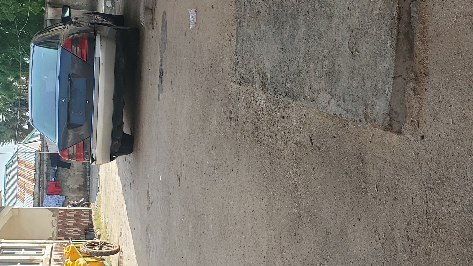 The image depicts an outdoor scene in Jalingo, Nigeria. In the foreground, a dirt and concrete path is visible, with some debris scattered on the ground. To the left, a yellow cart with a wheel is positioned near a building. In the background, a dark-colored sedan is parked, facing away from the viewer. Clothes are hanging on a line between two structures. The overall impression is one of a residential or informal area within the city.