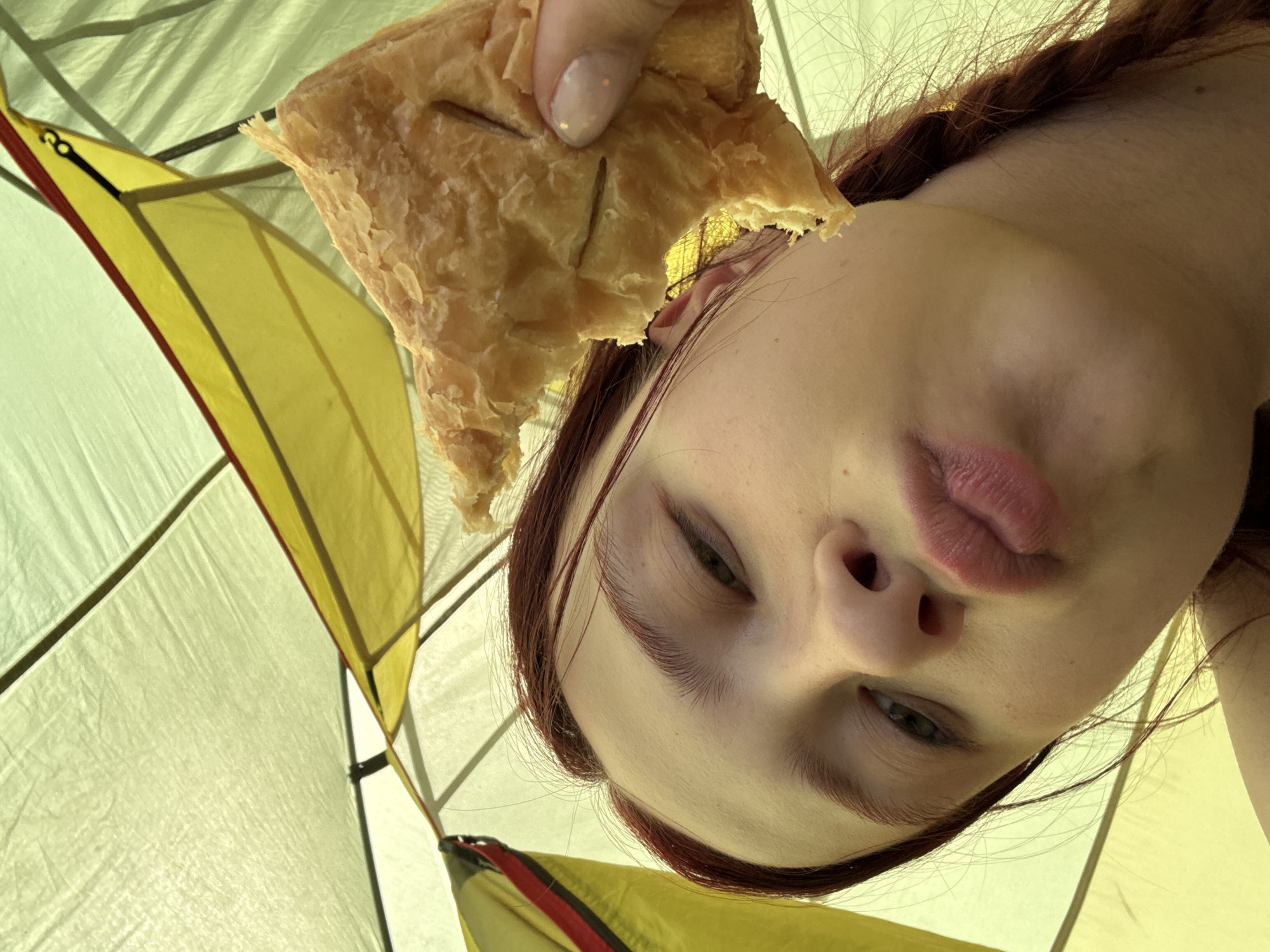 A close-up, low-angle shot looking up at a person inside a tent. The person is holding a partially eaten pastry. The tent has a yellow and white fabric interior, with a yellow pole with red trim. The person has fair skin, reddish-brown hair, and is looking down at the camera with a slight pout. Their hair is braided. The person's hand is holding the pastry near their face. No indication of location is present.