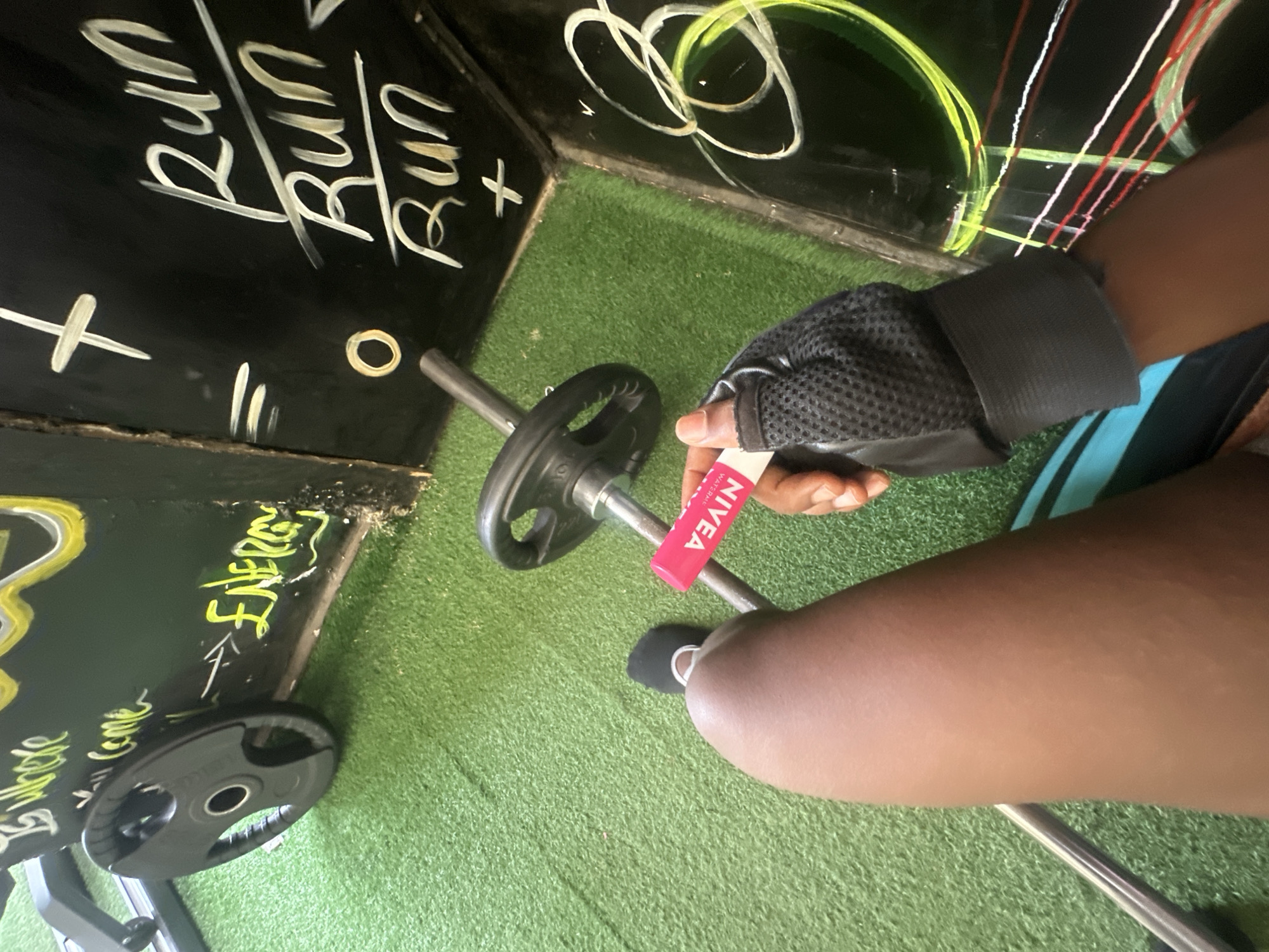A gym setting featuring artificial turf. A person wearing a gray weightlifting glove holds a pink Nivea Water Space lip balm next to a weight bar. Weights rest on the bar and on the floor. Writing in white and yellow adorns a black board. The words "Run" and "Energy" are visible. The location is Iddo Tudunwada, Nigeria.