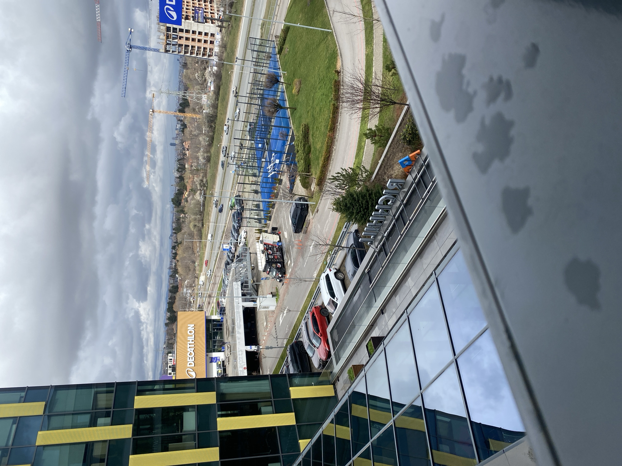 An elevated daytime view captures a section of Sofia, Bulgaria, with architectural and commercial elements predominating. The frame's foreground features sections of a contemporary building exhibiting a pattern of yellow and green tinted glass panels. Adjacent is a building with the signage for "DECATHLON" visibly displayed, indicating a retail establishment.
A road is partially visible, with multiple vehicles parked or moving along it, including a red car, a white car, and a dark-colored truck, among others. Behind the Decathlon building, blue-colored tennis courts or other sports facilities are partially observable.
In the backdrop, the cityscape is characterized by a mixture of residential or commercial buildings and some construction activity, including a crane. The sky is overcast with gray clouds. The image is taken from a high vantage point, possibly from a building, and the view is partially obscured by water droplets on the window or structure from which the photograph was captured.