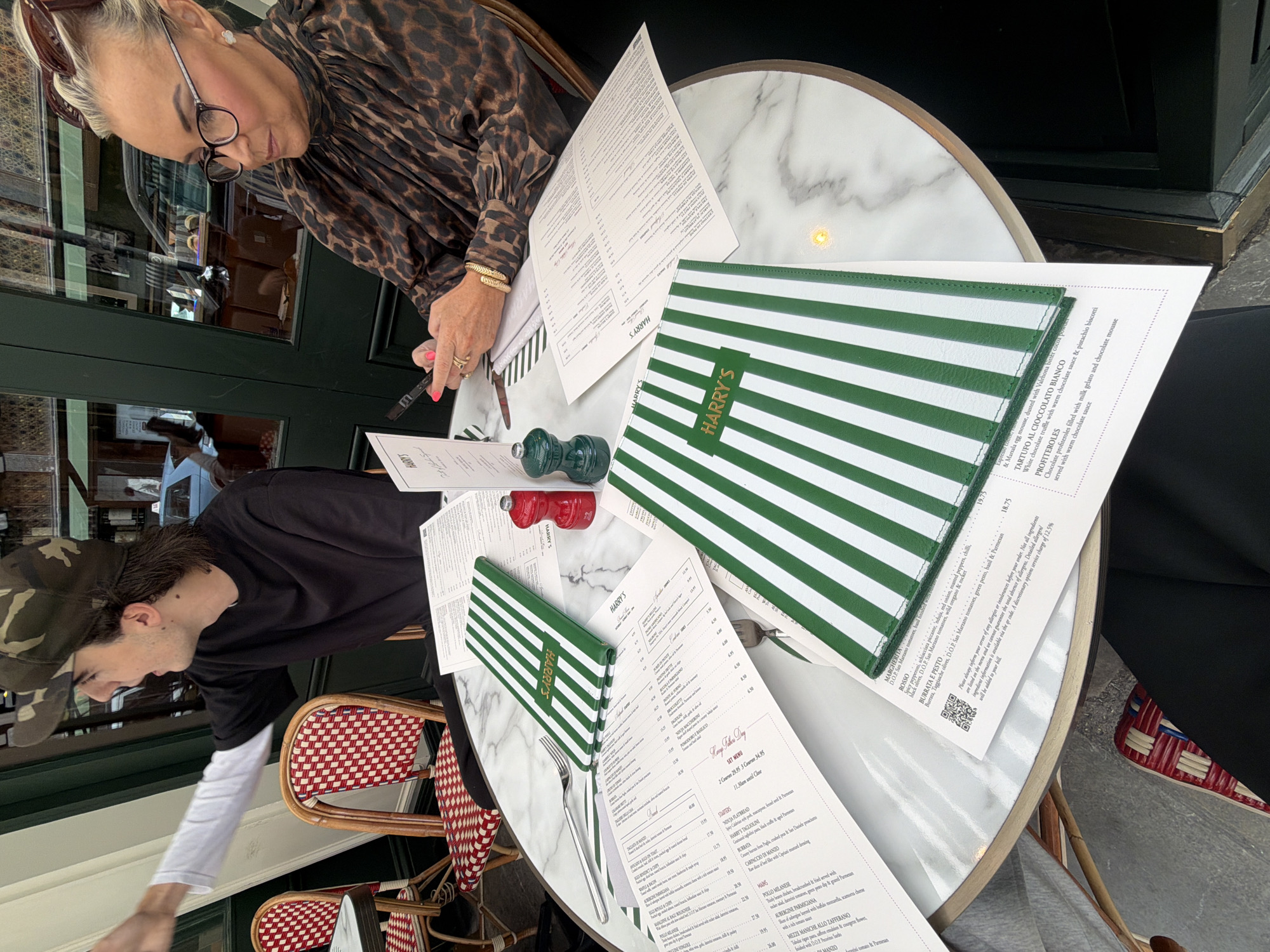 The image shows a table at what appears to be Harry's Bar, located in London, United Kingdom. Two people are seated at the table. On the left, a young man wearing a camouflage baseball cap and a black t-shirt with a white long-sleeved shirt underneath is seen from the side, reaching towards the camera. On the right, a woman with blonde hair styled up and wearing glasses and a leopard-print top is looking down at her phone, which she is holding in her hands. On the marble tabletop are several menus, two of which are green and white striped covers with "HARRY'S" written on them in gold. A red and a green salt and pepper grinder are also on the table, as well as cutlery. The setting appears to be outdoors, with a glimpse of a street and a building through a window in the background.