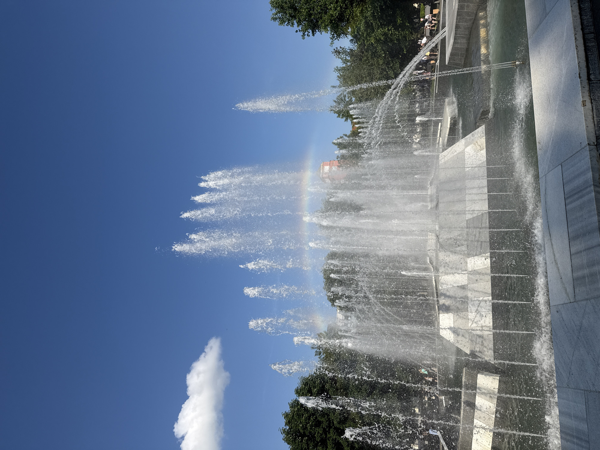 This image captures the Water Cascade Fountain in Pleven, Bulgaria, under a clear blue sky featuring a single white cumulus cloud in the upper left. The foreground is occupied by the large, multi-tiered fountain, constructed from light-colored stone with darker, contrasting sections, forming a series of basins and geometric shapes. Numerous powerful water jets are active, projecting water upwards and outwards, creating a dense mist and spray. A distinct, partial rainbow arc is visible within the central spray of the fountain. Behind the fountain, a dense line of mature green trees provides a natural backdrop. Partially visible through the trees and fountain mist on the right side is a reddish-orange building featuring a distinctive tower or spire structure. Several individuals are present in the mid-ground, walking, standing, or sitting near the fountain and along the treeline, dressed in casual attire. The scene is brightly illuminated by natural daylight.