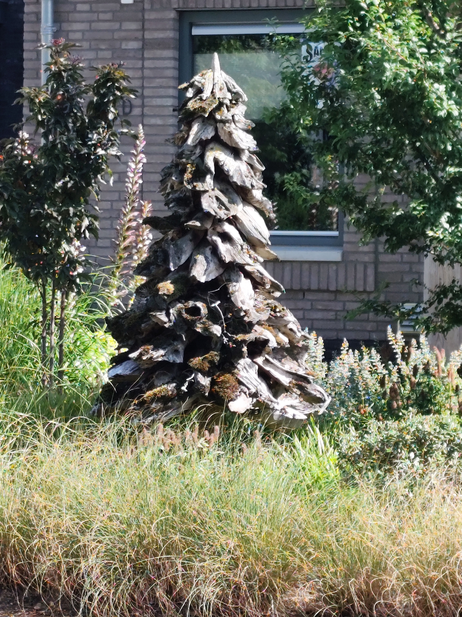 The image shows an outdoor decorative garden scene. In the center foreground, a conical sculpture made of weathered wood is prominently displayed. The sculpture is roughly textured and appears to be composed of stacked, overlapping pieces of wood. Surrounding the sculpture is lush, green grass and various other plants. In the background, a brick building with a large window is visible. The window reflects the surrounding greenery and a partially visible sign. To the left of the sculpture, a small tree with dark green leaves and some red berries can be seen. To the right of the sculpture, a cluster of tall, slender plants with light purple flowers are growing. The overall impression is of a well-maintained garden with a unique artistic element.