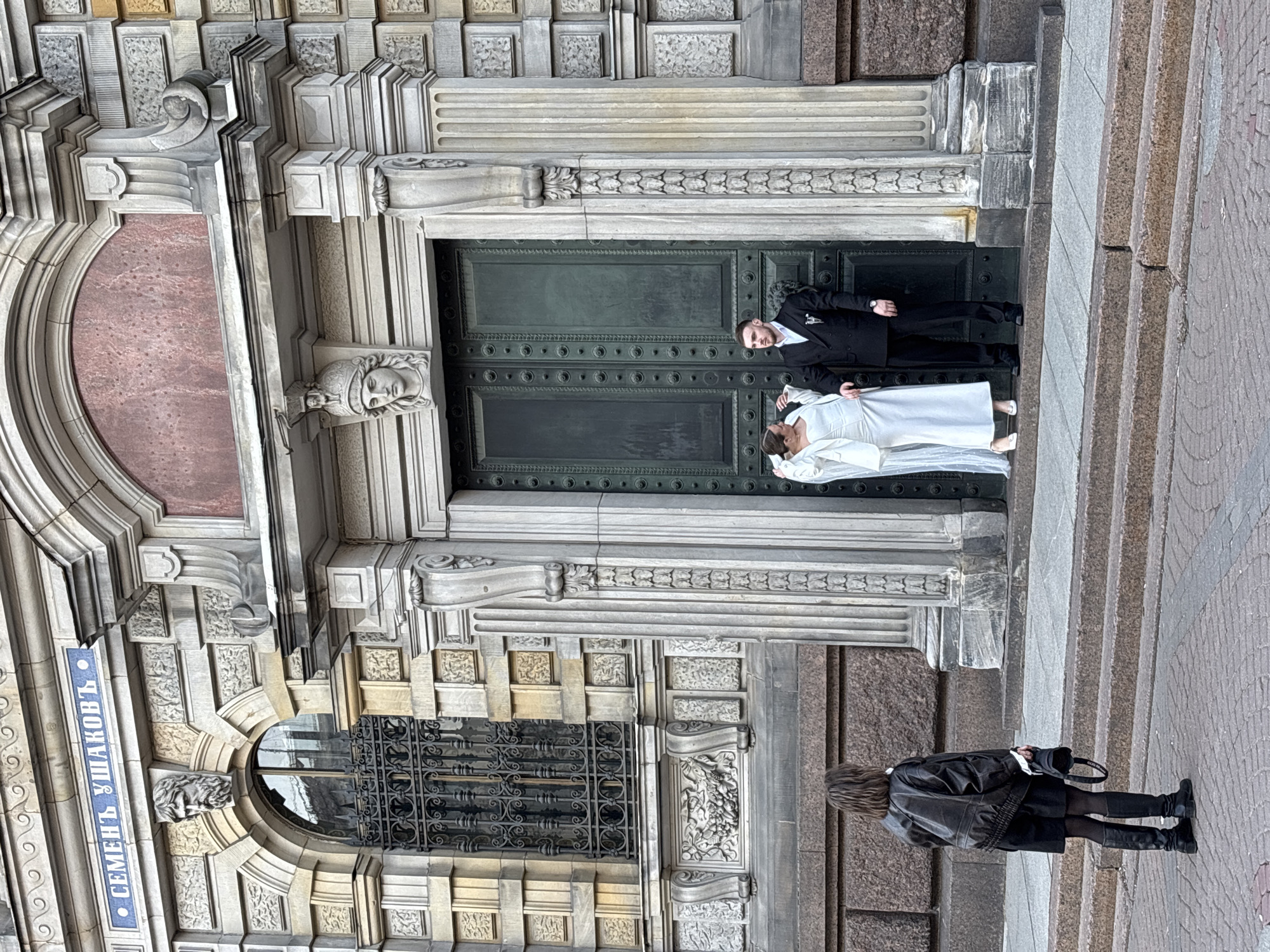 A street scene in Saint Petersburg, Russia, shows a wedding photo shoot outside a building with ornate architectural details. A bride in a white dress and a groom in a black suit lie horizontally in the doorway of the building. The building has a plaque reading "СЕМЕНЪ УШАКОВЪ" (Semyon Ushakov), possibly an indicator of the building's historical association with the artist. A person in a black leather jacket, black dress, and boots stands on the pavement in front of the building, holding a black purse. The architecture is characterized by intricate carvings, a stone facade, and a large green door.