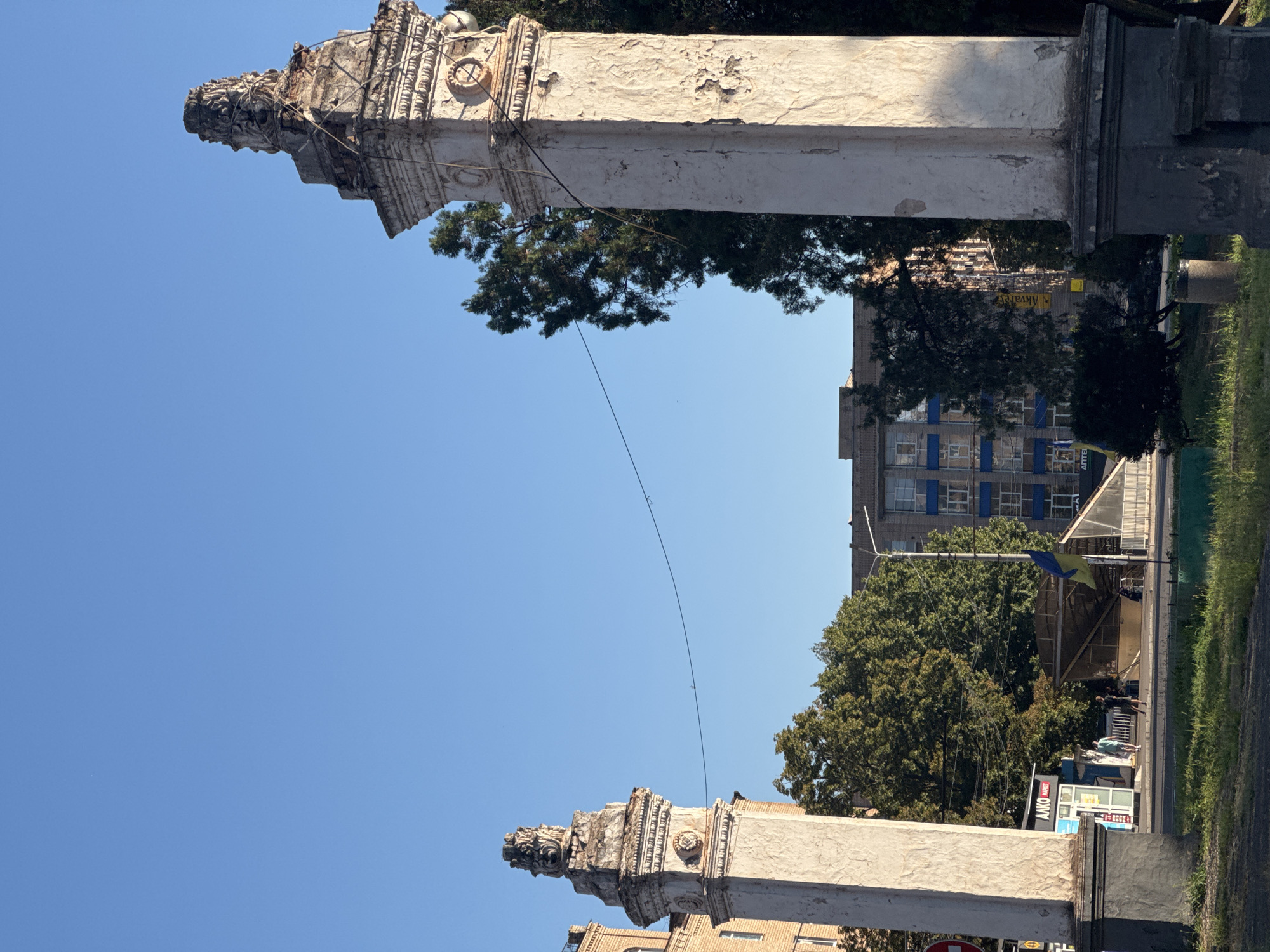 The image captures an outdoor scene with two ornate, weathered stone pillars flanking the foreground. The pillars are tall and rectangular with decorative capitals topped by sculpted elements that appear to be heads or animalistic figures. Both pillars show signs of age and deterioration, with chipped paint and rough textures. A thin wire stretches horizontally between the tops of the pillars. In the background, a clear blue sky dominates the upper portion of the frame. Below the sky, a line of mature green trees partially obscures a multi-story building with a blue and white facade. To the left of the pillars, a brick building is visible. On the ground level, there is a paved area with some grass. Several people are walking on the paved area, and a yellow and blue Ukrainian flag is visible, attached to a pole. A small kiosk with signage in Cyrillic is also present. The scene appears to be a public space in the city of Kryvyi Rih, Ukraine, possibly an entrance or monument.