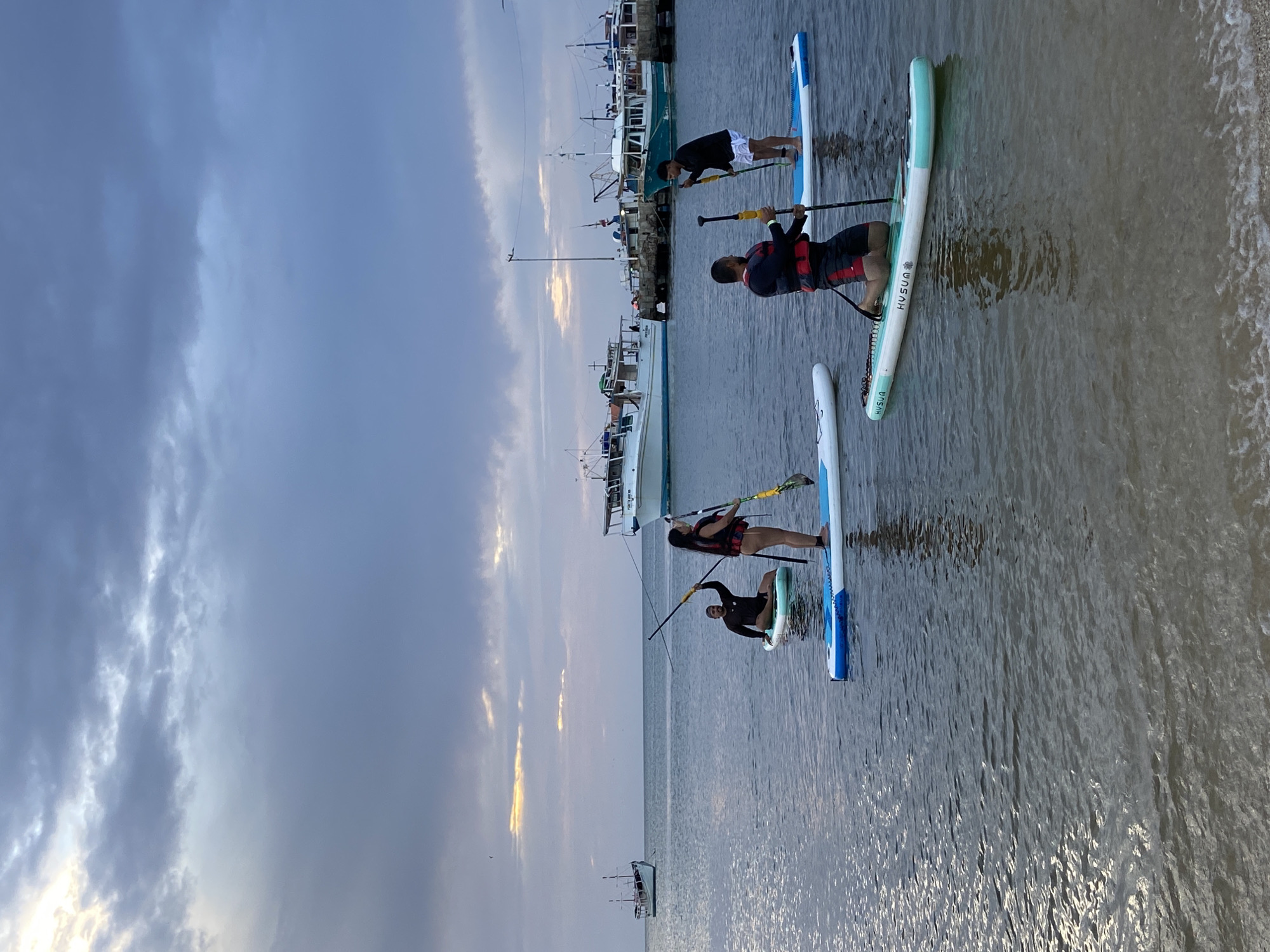 The image depicts a group of four individuals paddleboarding in the waters of Juan Griego, Venezuela. The sky is overcast with hints of sunlight breaking through the clouds. In the background, several boats are docked near a pier, with one larger white boat prominently positioned.
The paddleboarders are distributed across the water. One individual, wearing a life vest, is kneeling on a blue and white paddleboard, holding a paddle. Another person, also in a life vest, is standing on a similar paddleboard and is in the process of paddling. A third individual, in a black long-sleeved shirt and white shorts, stands on a paddleboard, holding a paddle. The fourth person is further in the distance, kneeling on a paddleboard. The water is calm with gentle ripples, and the shore is visible in the foreground. The overall atmosphere suggests a leisurely activity in a coastal setting.