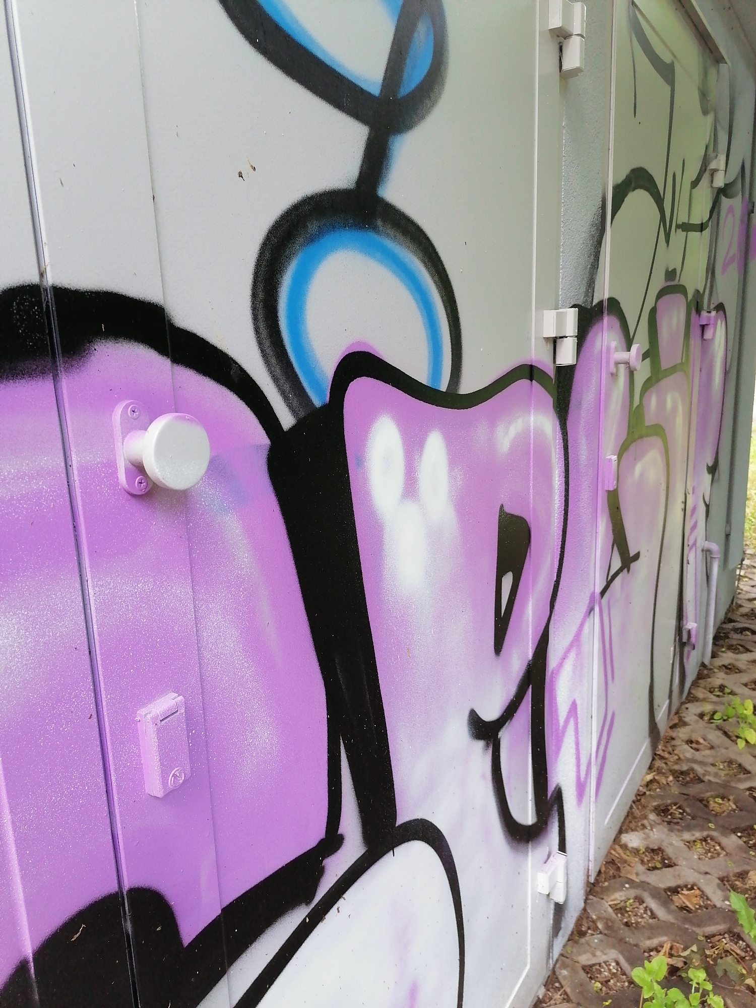 The image shows a close-up, angled view of a gray metal utility box covered in graffiti in Unterhaching, Germany. The graffiti features large, stylized letters in purple, black, and white, with blue outlines on some elements. Two white door handles or knobs are visible on the left side of the box, one on a raised panel. White hinges are also visible where the panels of the box meet. To the right of the box, a section of patterned concrete pavers with some sparse green vegetation is visible. The overall impression is one of urban decay or artistic vandalism in an outdoor setting.