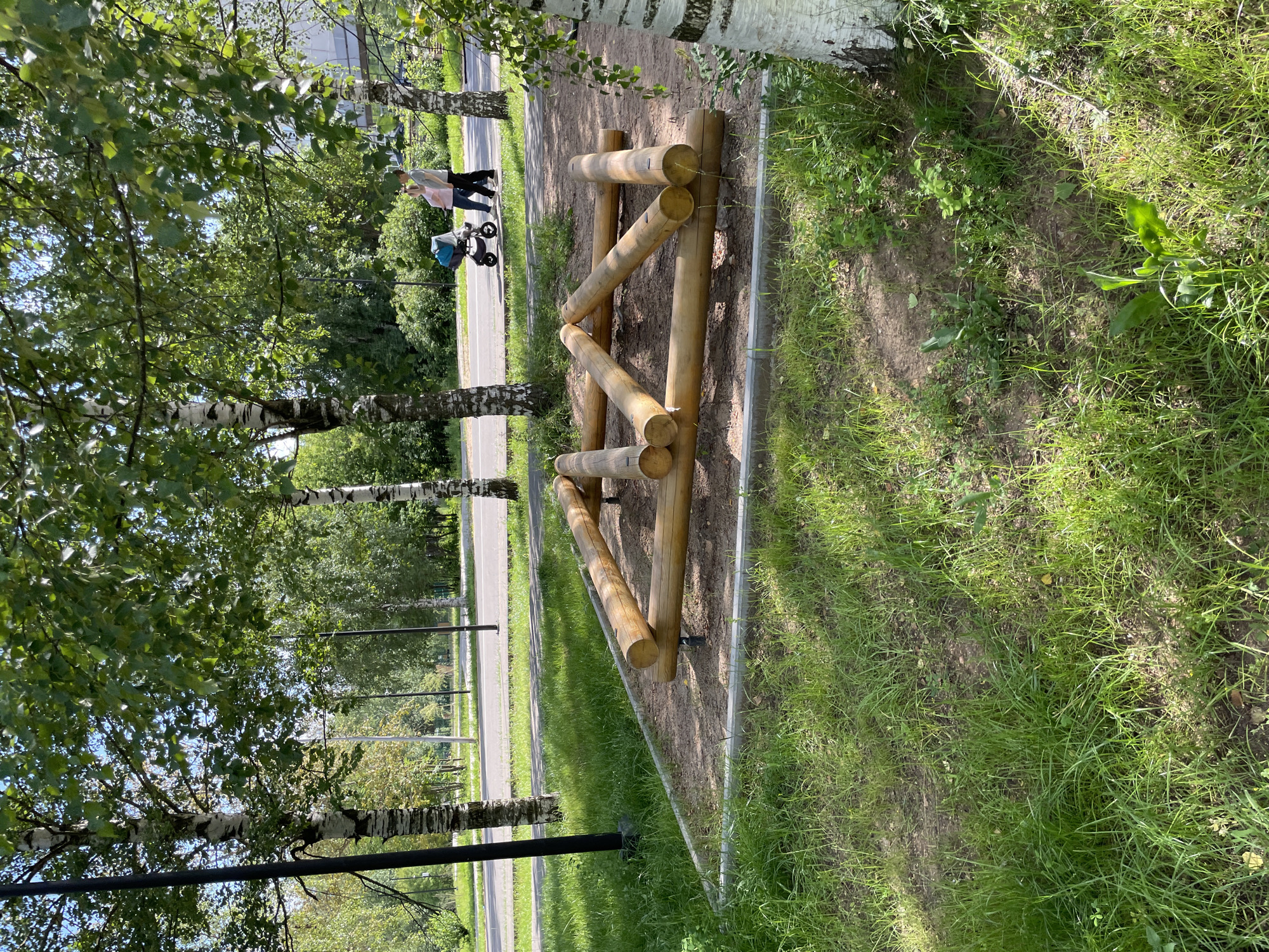 In Kirovo-Chepetsk, Russia, an outdoor fitness obstacle made of wooden logs is situated on a sand-covered area surrounded by grass and bordered by a concrete curb. The obstacle is a triangular structure formed by several logs. In the background, two people are walking on a paved path next to a street. One person is pushing a stroller. The scene is framed by birch trees with dappled sunlight filtering through their leaves.