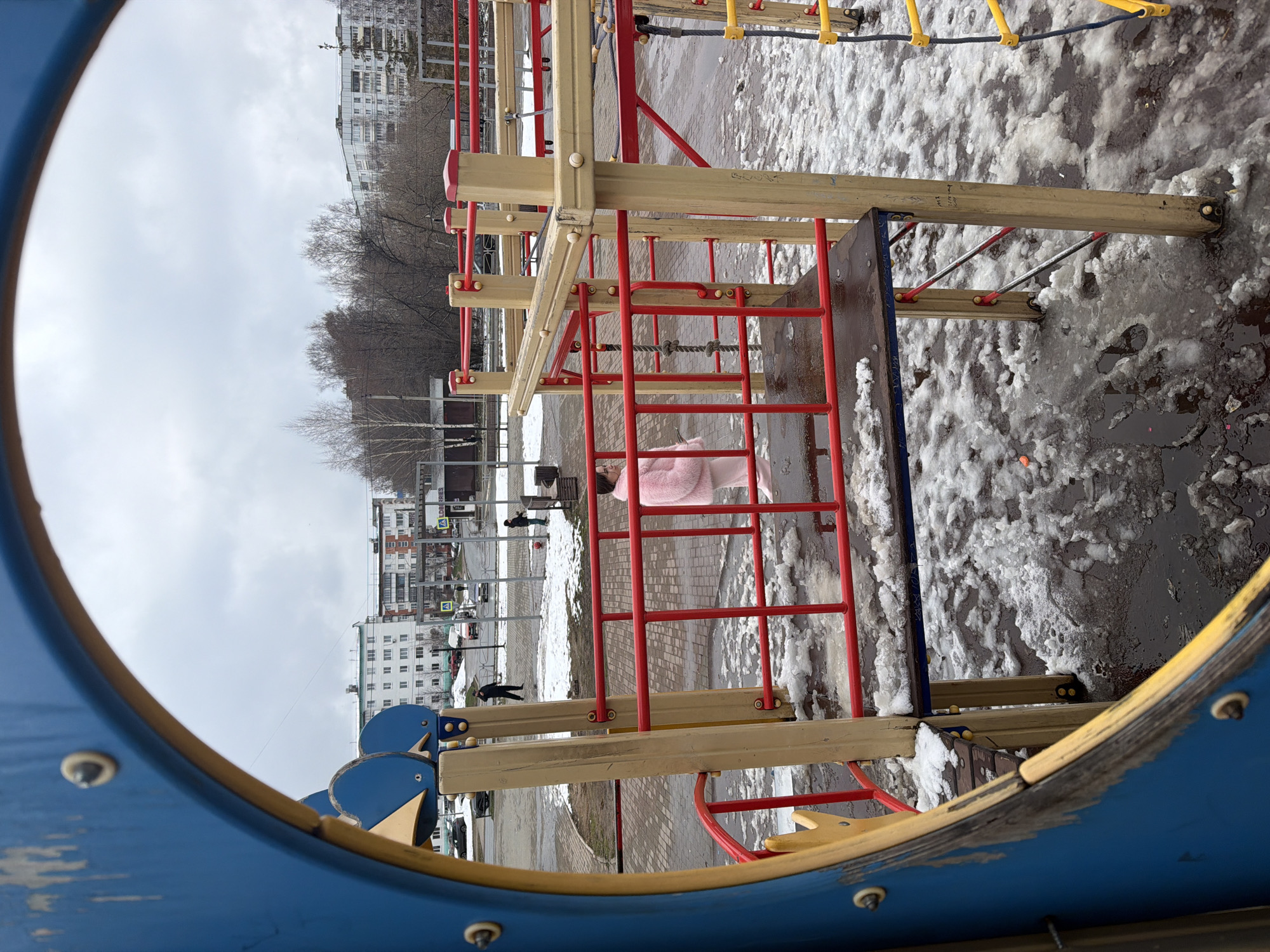 Observed from within the blue and yellow frame of a children's playground structure, the image captures a late-winter or early-spring scene in a residential area of Chaikovsky, Russia. Patches of melting snow and puddles are visible on the ground. Several buildings line the background, suggesting a densely populated urban environment. Bare trees and overcast skies denote the season. Within the playground structure, a red and tan ladder is the most prominent element. A child in a pink coat is visible in the background near the playground. Several adults are walking on a sidewalk, including a man in a dark coat and a man wearing a red jacket. Other elements include traffic lights, cars, sidewalks, and buildings in the background.