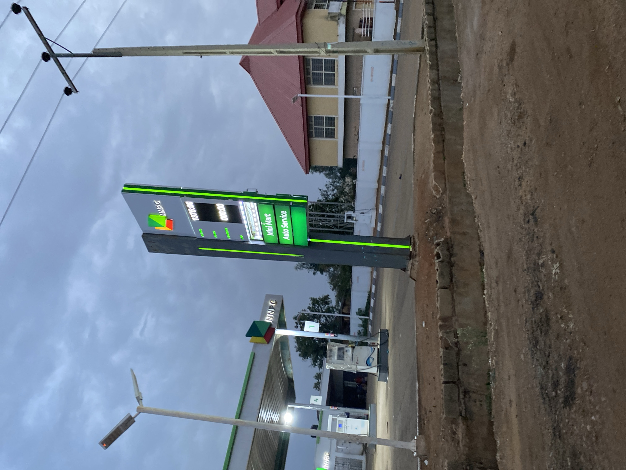 The image depicts a gas station in Kuru Dungus, Nigeria. A sign displays the price of petrol as 1375:00. The gas station also offers "Mini Mart" and "Auto Service". Several gas pumps are visible. The sky is overcast. The ground is dirt and pavement. There are utility poles with lights. A building with a red roof is in the background.