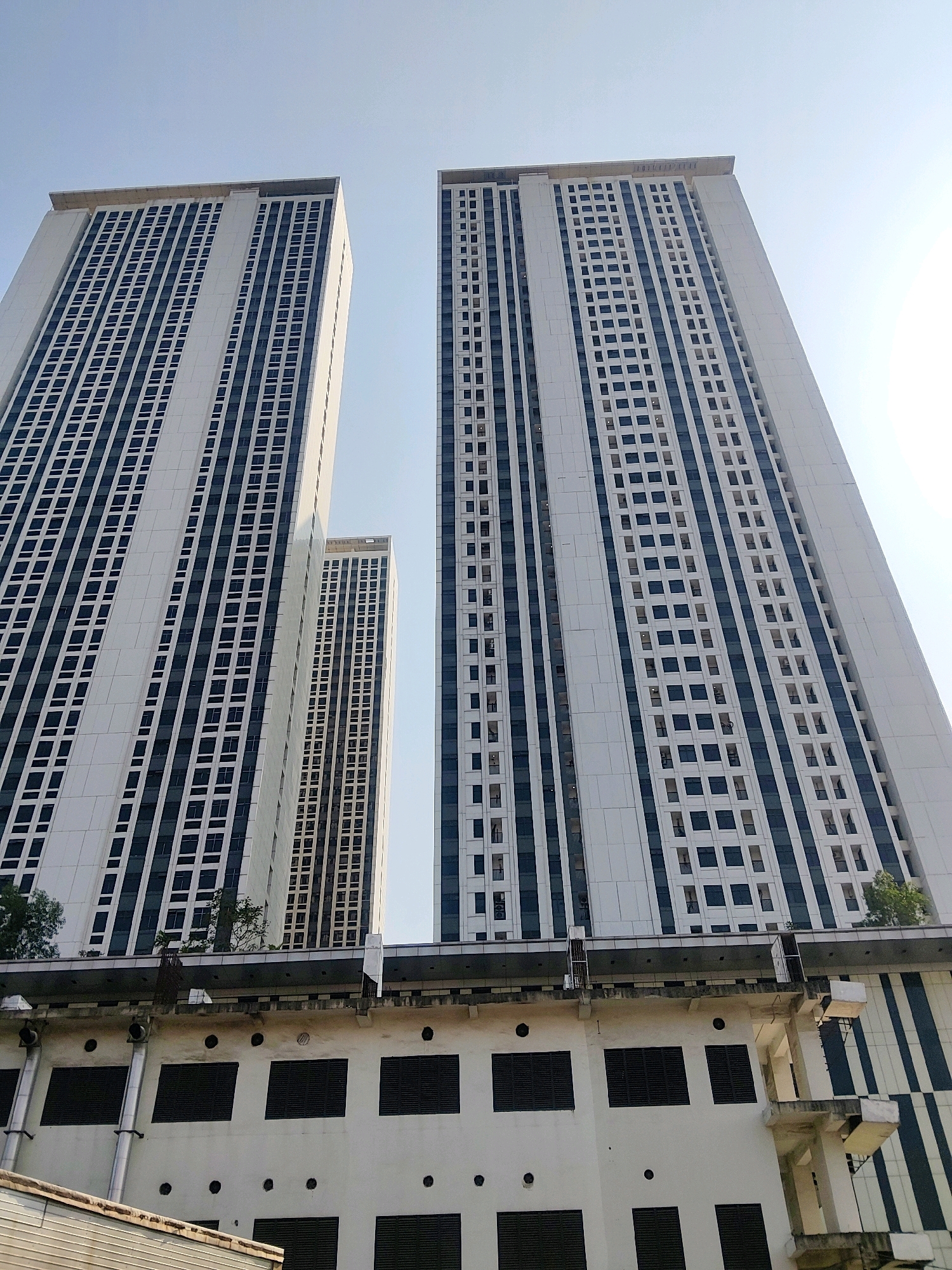 The image shows three high-rise apartment buildings in Sukaresmi, Indonesia. The buildings are tall and rectangular, with white exteriors and dark blue or black windows. The windows are arranged in a grid pattern on the facades of the buildings. The buildings are viewed from a low angle, looking up towards the sky. The sky is clear and blue, with some sunlight visible. The foreground of the image shows a lower building with a more industrial appearance, featuring exposed pipes, vents, and dark rectangular windows. The overall scene depicts a dense urban environment with modern residential structures.