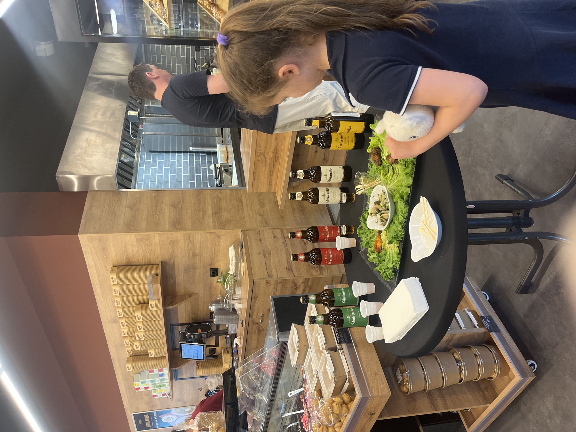 In Belgrade, Serbia, a girl with a purple hair tie, wearing a dark blue shirt, stands at a circular table, holding a small, white, stuffed animal. She is looking at a spread on the table, including bottles of Valdermuzia beer in yellow, brown, and green labels, small plastic cups, and plates of food with lettuce and a white sauce. An adult male in a black shirt is also at the table. The table has a black top and metal legs, with a wooden shelf below displaying cardboard containers. To the left, there's a display case with pastries and sweets, and further back, a wooden wall with stacks of disposable coffee cups. A stainless steel kitchen area is visible in the background.