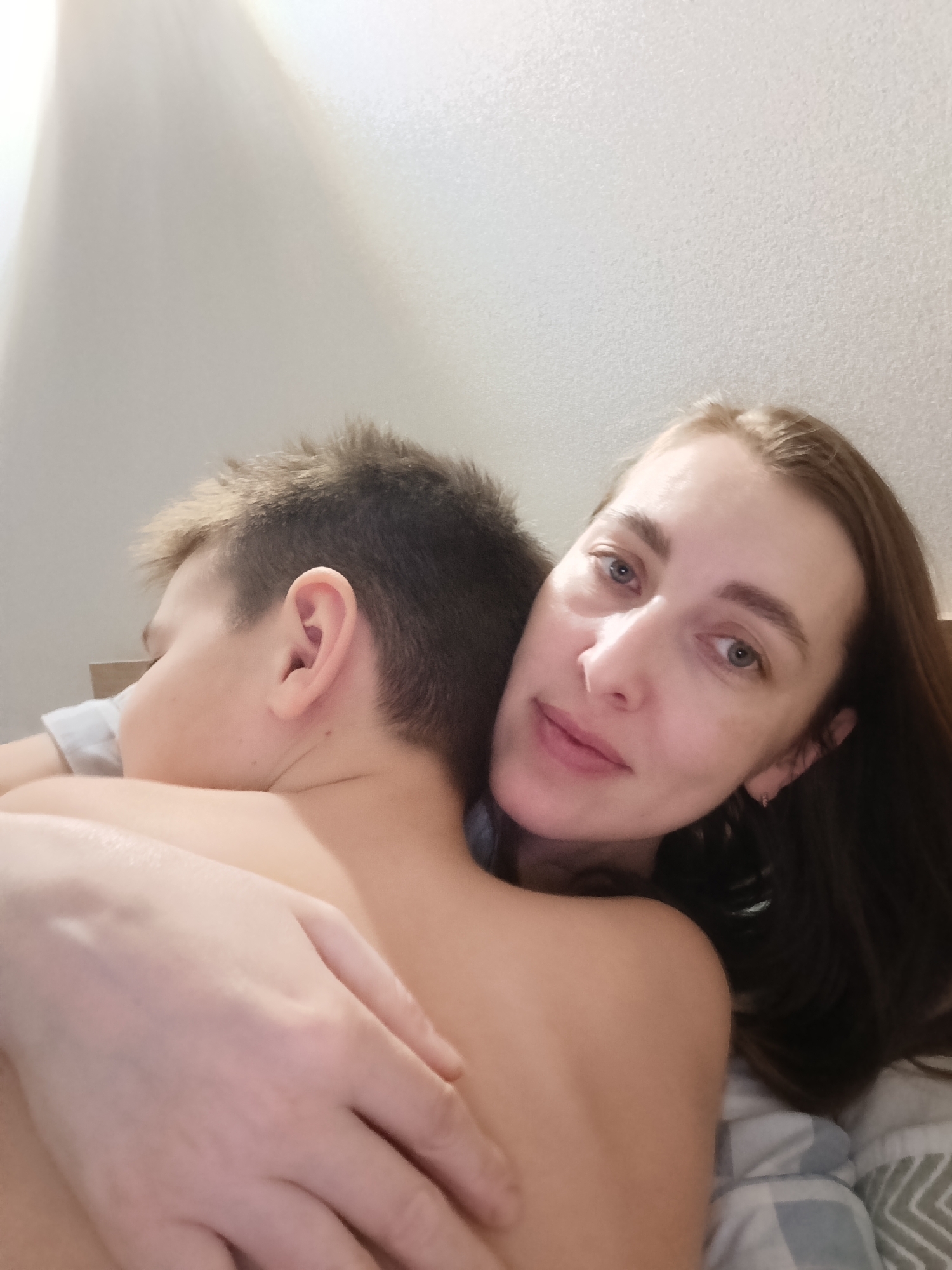 The image shows a woman embracing a young boy. The woman is looking at the camera with a slight smile. The boy is turned away from the camera, with his face resting against the woman's chest. Both are lying on a bed covered with a grey and white checkered blanket. The background is a plain white wall. There is no indication of a specific location or city.