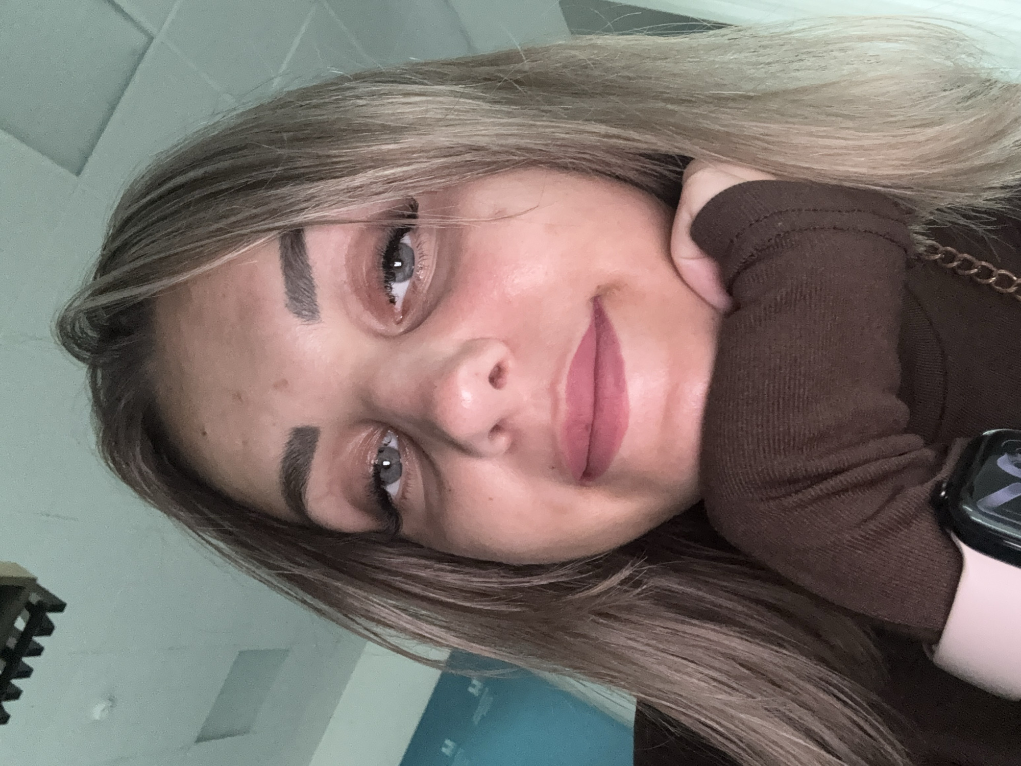 A young Caucasian female with blonde hair is resting her chin on her hand, looking at the camera. She is wearing a brown turtleneck sweater. A smartwatch is visible on her left wrist. The background consists of a white ceiling with visible lighting fixtures and a blue wall. The scene appears to be indoors, possibly an office or public space. The image was captured in Yuzhno-Sakhalinsk, Russia.