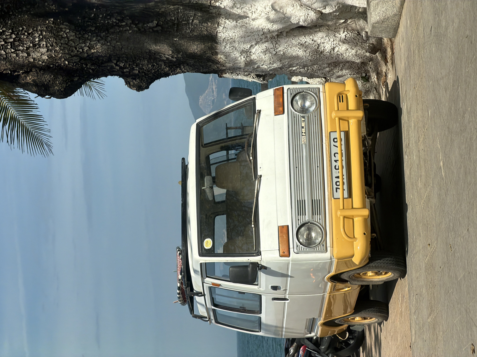 A white and yellow Daihatsu Hijet van is parked on a paved surface near the ocean in Nha Trang Ward, Vietnam. The van has a surfboard strapped to its roof rack. A large tree with a whitewashed trunk stands to the right of the van. In the background, the ocean and distant mountains are visible under a partly cloudy blue sky. The van's license plate is visible and reads "70A-012.10". A motorcycle is partially visible on the left side of the image, parked next to the van.
