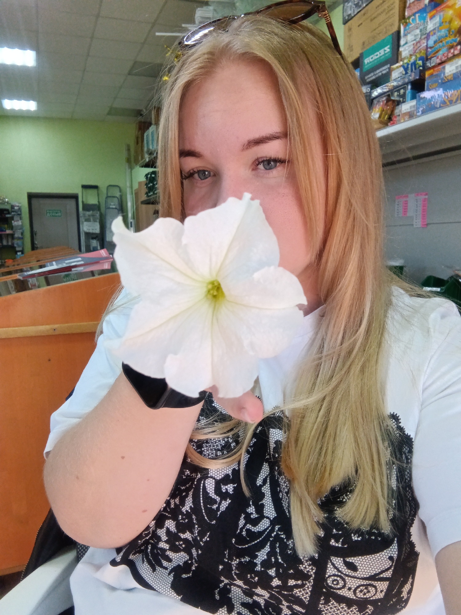 A woman with blonde hair is holding a white flower to her face in a room with shelves stocked with items. She is wearing a white shirt with a black lace pattern. The background suggests a retail or storage environment, possibly a shop or warehouse. There is a door with a green exit sign visible in the left background. The lighting is fluorescent and consistent with an indoor setting. The image appears to have been taken in the city of Fokino, Russia, based on the provided context.