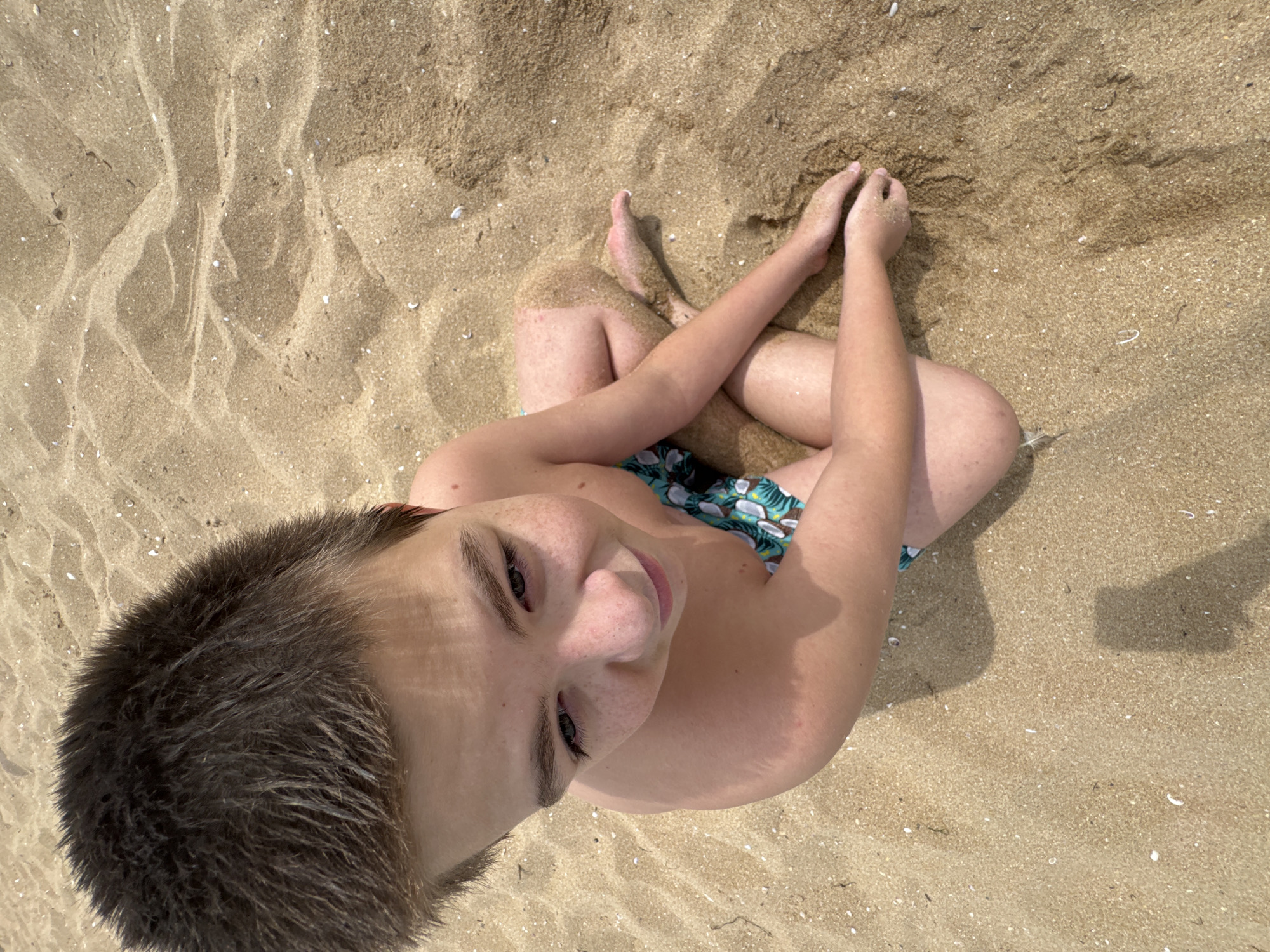 A young boy is sitting cross-legged on a sandy beach, his body angled slightly towards the right of the frame. He is looking downwards and to his left, with a slight smile on his face. The boy has short, dark brown hair and freckles across his nose and cheeks. He is wearing swim trunks with a tropical print. The sand around him is light brown and appears slightly damp in places, with small shells scattered throughout. The lighting suggests it is daytime, with shadows cast by the boy's body indicating the sun is overhead. The scene is set on a beach, likely in Sozopol, Bulgaria, a popular Black Sea resort known for its sandy coastlines.