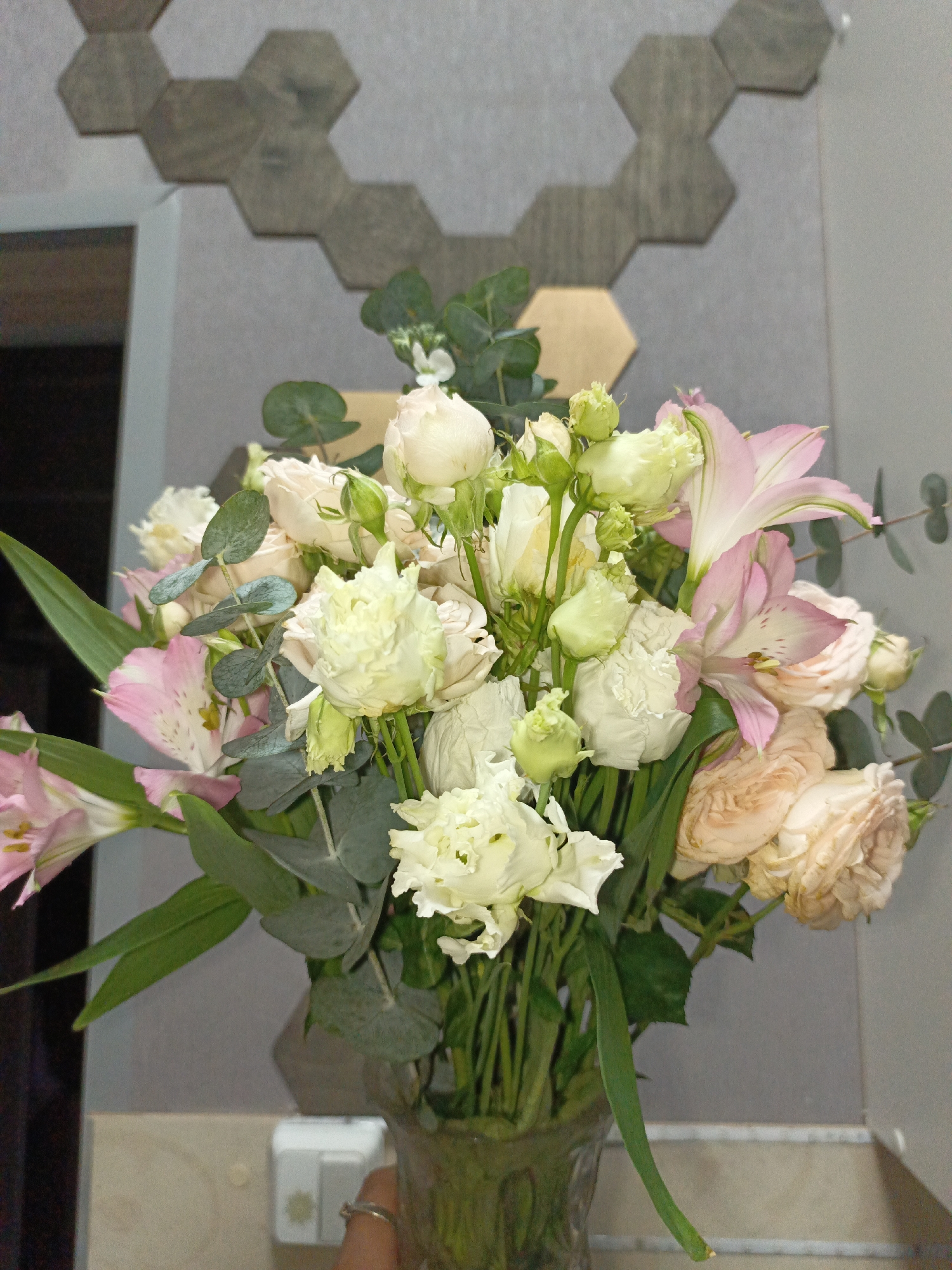 A bouquet of flowers in a vase is set against a grey wall decorated with dark grey and gold hexagonal tiles. The bouquet contains white and pale peach roses, white lisianthus, pink alstroemeria lilies, and eucalyptus foliage. The vase is dark green glass, and a hand wearing a ring is visible holding it. A light-colored counter is beneath the vase. In the background, to the left of the flowers, is a partially visible door frame or corner of the room. An electrical outlet is visible on the wall beneath the flowers. The location is Марковский, Russia.