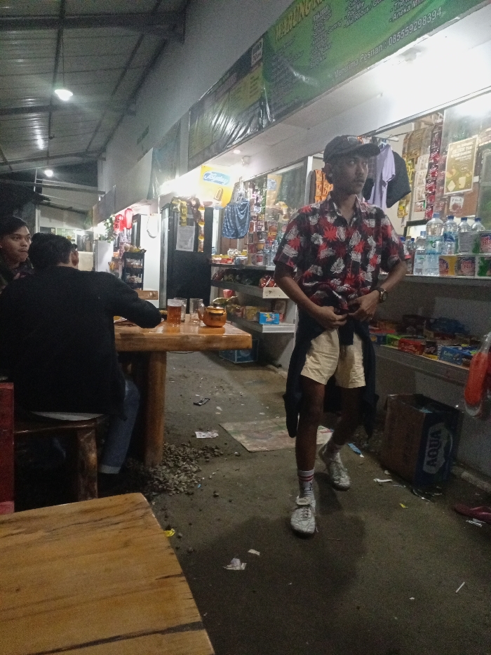 This image captures a nighttime scene at a modest, open-air or semi-enclosed "warung" (small shop or food stall) in Pangalengan, Indonesia. The area is illuminated by artificial fluorescent lights, creating distinct shadows.
In the foreground and to the right, a young man stands, slightly angled towards the left. He is wearing a dark cap, a short-sleeved shirt with a red, black, and white leaf or floral pattern, light-colored shorts, and white sneakers with striped socks. A dark garment is tied around his waist, and he is holding the hem of his shirt with both hands, seemingly adjusting it.
To the left, two men are seated at a rustic wooden table made of rough-hewn timber. One man, seen from behind, wears a dark long-sleeved shirt and blue jeans, sitting on a wooden stool. A glass containing an amber liquid and a small bronze-colored teapot are on the table in front of him. Another man is partially visible to the left of the seated man. They appear to be engaged in casual conversation or resting.
The background reveals the interior of the warung, featuring shelves stocked with various packaged goods, snacks, and bottled drinks, including prominently displayed "Aqua" brand water bottles. A blue "AQUA" cardboard box is visible on the floor near the standing man. Above the shelves, banners and signs are displayed; visible text includes "WARUNGKA..." (likely part of the shop's name), "menerima Pesanan" (receives orders), and a phone number "085559298394". The floor is a mix of concrete and dirt, with small pebbles, discarded paper, and other debris scattered around, suggesting a casual, well-used establishment. The overall scene depicts a relaxed, local social setting in the evening.
