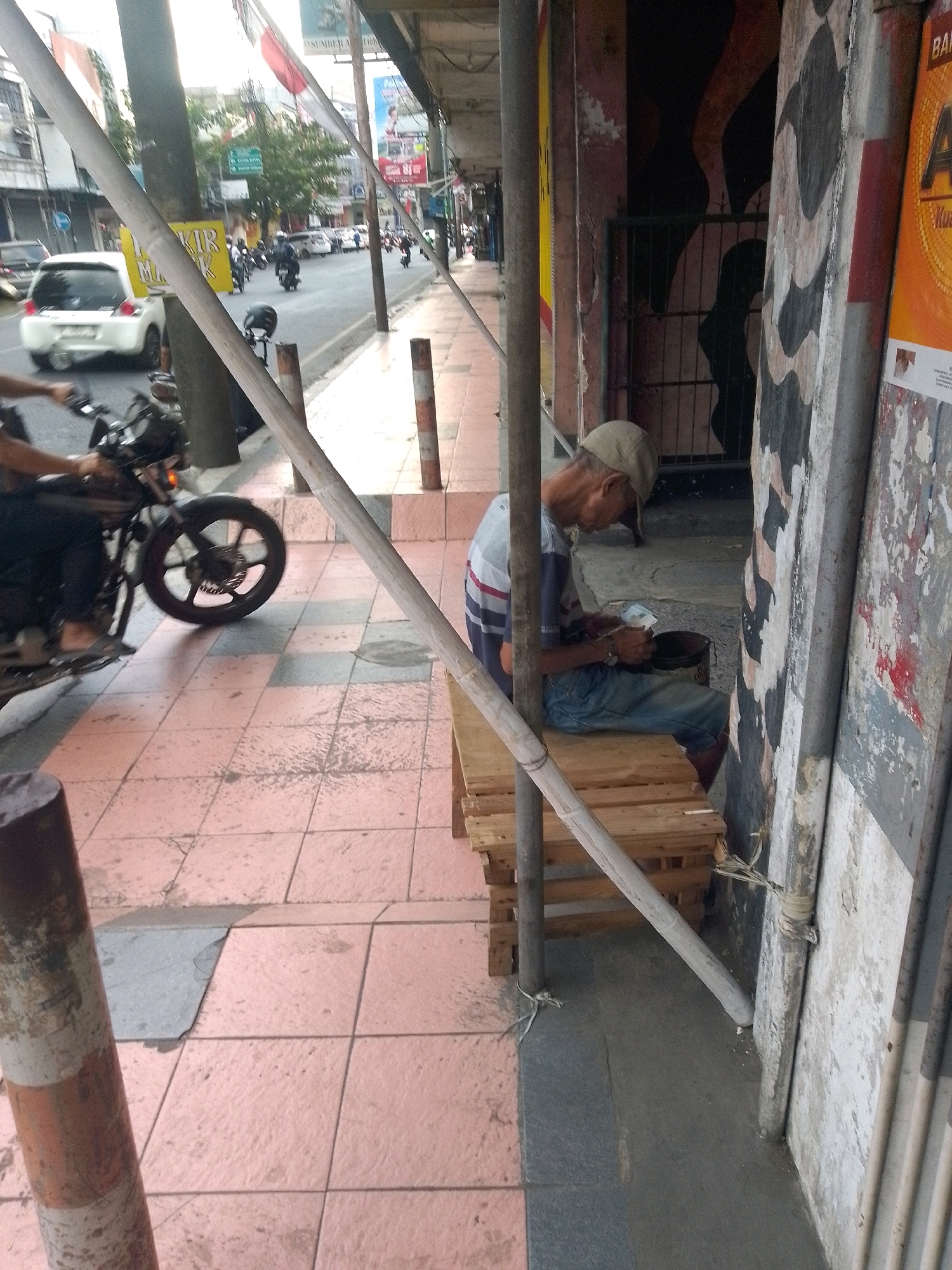 The image depicts a street scene in Jember, Indonesia. In the foreground, a man wearing a light-colored cap, a striped shirt, and blue jeans is seated on a wooden pallet structure. He is holding some papers in his hands and appears to be interacting with a dark-colored bucket beside him. The man is positioned near a building with a weathered facade. To the left of the frame, a motorcycle is partially visible, with its front wheel and handlebars in view. Beyond the seated man and the motorcycle, the street is visible, with a white car parked on the side and other vehicles and buildings further in the background. The street is paved with pinkish tiles. Overhead, scaffolding and poles are present, suggesting ongoing construction or temporary structures. Several flags, including what appears to be the Indonesian flag, are visible in the background, fluttering in the breeze. The overall environment suggests a busy urban setting.