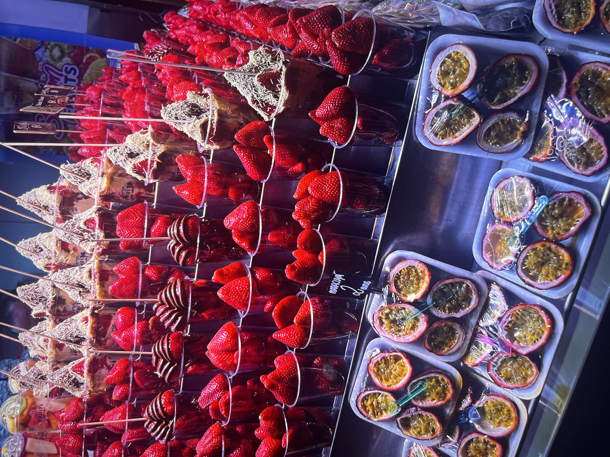 The image shows a display of fruits, likely for sale, in a brightly lit indoor setting, possibly in Unknown, Unknown. The main focus is on a variety of strawberries prepared in different ways: some plain in clear plastic cups, some with chocolate drizzled on them, and some on sticks covered in what appears to be white chocolate and sprinkles. To the right of the strawberries are containers of halved passion fruits, wrapped in plastic wrap. A small sign with text "maracuyá 2 euros" is visible next to the strawberries.