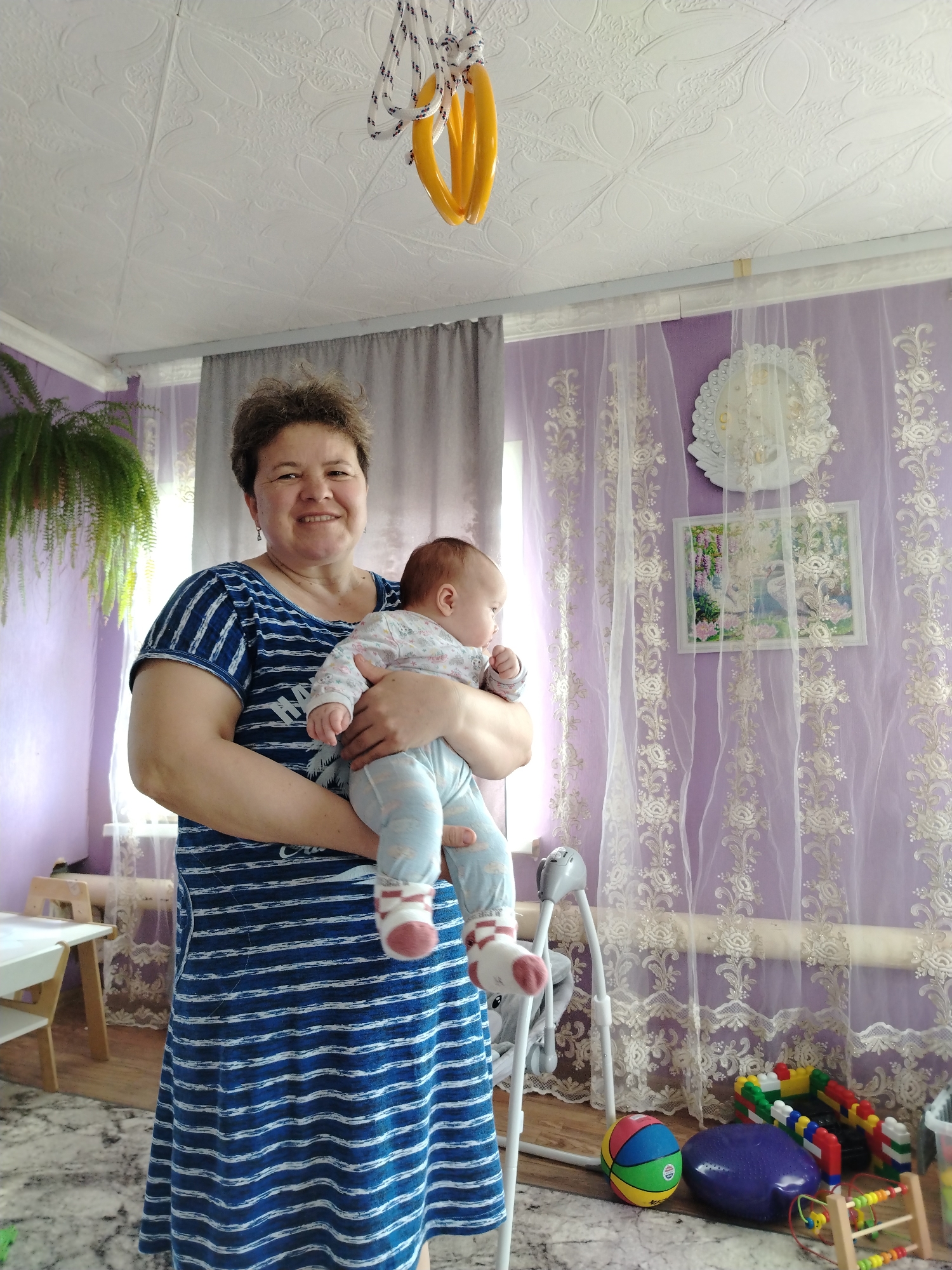 Here is a forensic description of the image:
**Scene:**
The photograph appears to be taken inside a home, likely a living room or nursery, in Гожан, Russia. The room has light purple walls with decorative lace trim and sheer curtains. A fern hangs from the ceiling in the left corner. The ceiling is textured with a floral pattern, and a set of yellow gymnastic rings hangs by ropes.
**People:**
A middle-aged woman is holding a baby. The woman is wearing a blue and white striped dress. She has short, dark hair and is smiling at the camera. The baby is wearing a patterned onesie with light blue pants and white socks with red accents.
**Objects:**
* **Furniture:** A small white table with chairs is visible in the background on the left side of the frame. A baby swing is located near the right.
* **Decor:** In addition to the curtains and wall trim, there is a decorative white wreath on the wall and a framed picture.
* **Toys:** A small collection of toys is located on the floor near the baby swing, including a ball, building blocks, and a purple exercise ball.
**Actions/Interactions:**
The woman is holding the baby in her arms. The baby appears to be looking around the room.
**Environment:**
The room has a warm, domestic feel with personal touches. The presence of the baby swing and toys suggests that the room is used or intended for child care.