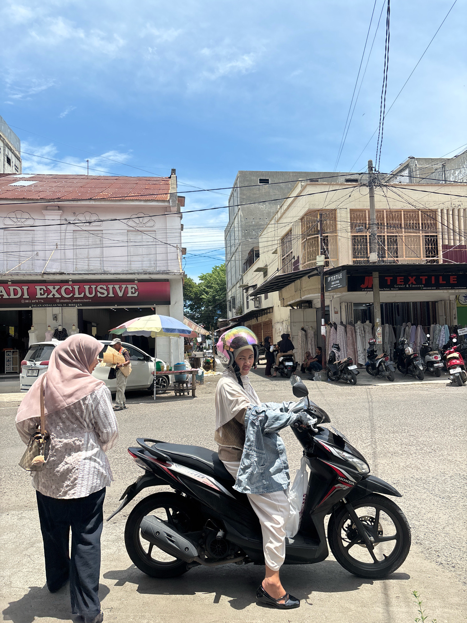 The image depicts a street scene in Bireuen, Indonesia, on a sunny day with a partly cloudy sky. In the foreground, a person wearing a light-colored hijab and a patterned blouse walks away from the camera, carrying a shoulder bag. To the right, a woman on a black Honda scooter, wearing a rainbow-colored helmet and a light-colored hijab, holds a denim jacket over her arm. She is looking towards the camera and appears to be smiling.
The scooter is parked on the side of the road in front of a row of shops. One shop has a prominent sign reading "ADI EXCLUSIVE" with "JALAN ANDALAS NO. 33 - BIREUEN" below it. Another shop to the right has a sign for "JP TEXTILE" and "JL. Andalas". Displayed outside the textile shop are various fabrics and mannequins. Several other scooters are parked along the street. In the background, buildings with white and cream facades line the street, some featuring ornate window grills. A white car is parked in front of the "ADI EXCLUSIVE" store, and a vendor is seen standing near a cart with a colorful umbrella. Utility poles and a dense network of electrical wires are visible overhead. The road surface is asphalt, and there are shadows cast on the ground.