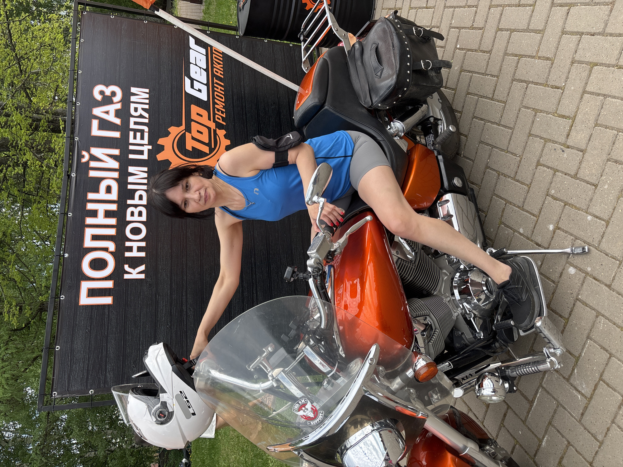 A woman wearing a blue tank top and grey shorts is posing on an orange motorcycle. She has dark hair and is smiling at the camera. The motorcycle is parked on a brick surface. Behind her is a black banner with orange and white text in what appears to be Russian. The banner also has a logo with the words "Top Gear" and a gear icon. To the left of the motorcycle, on the ground, is a white motorcycle helmet. The background includes trees and grass, suggesting an outdoor location.
